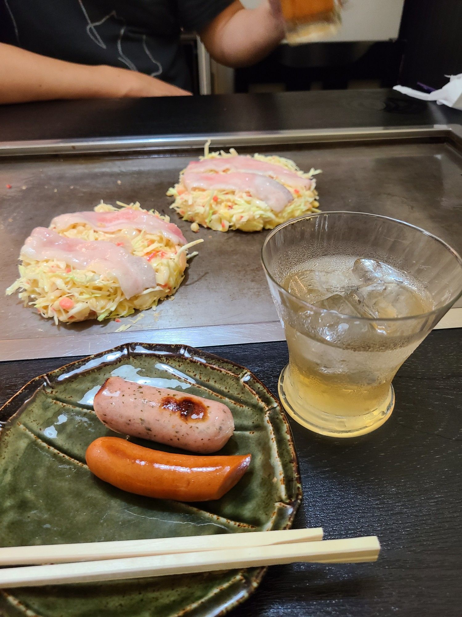 Two okonomiyaki sitting on a hot plate in the background with a small plate of sausages and glass of umeshu on the rocks in foreground