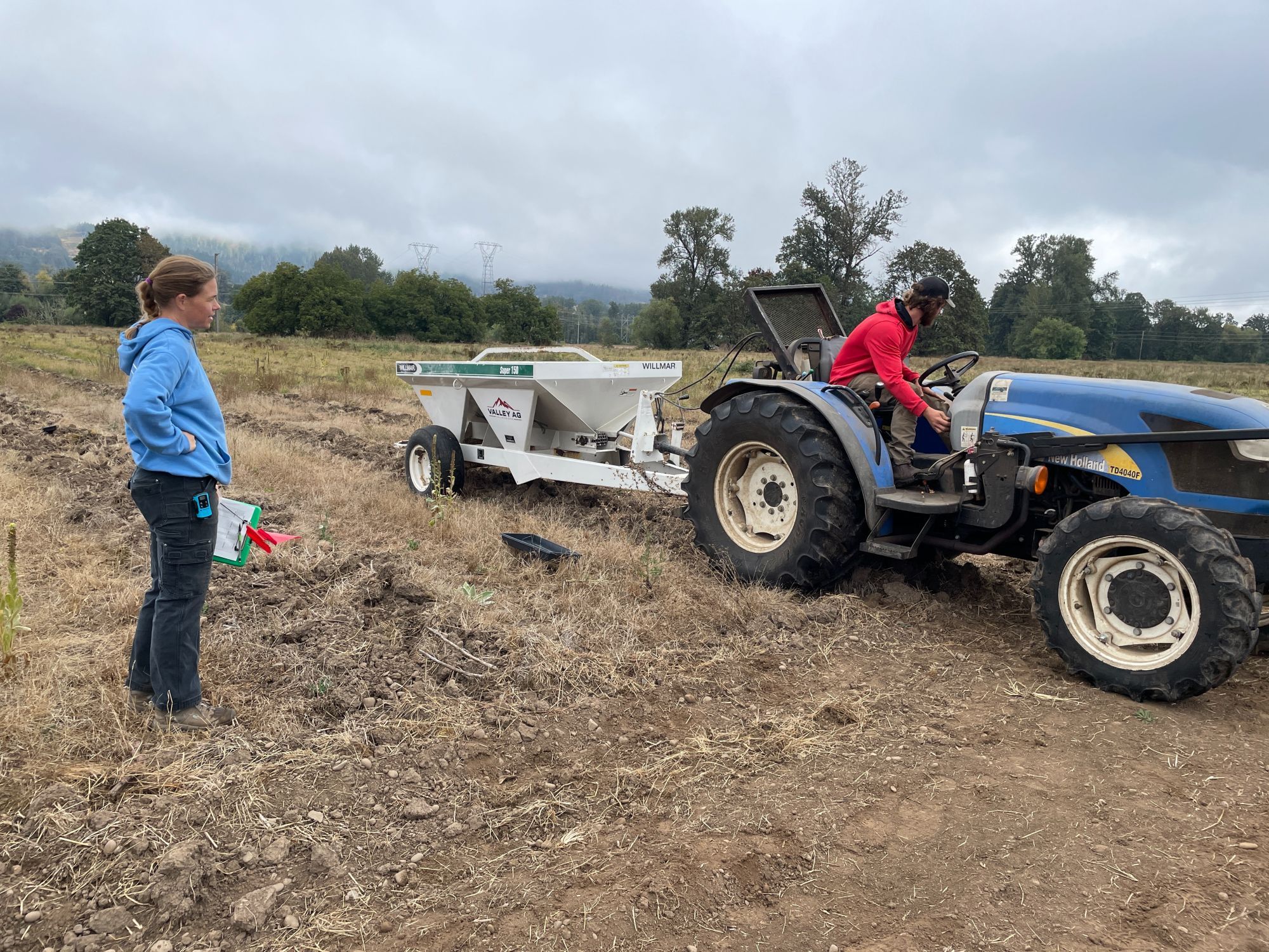 Emily supervises a tractor and spreader rig that's preparing to apply ground basalt dust to an open field ringed with trees.