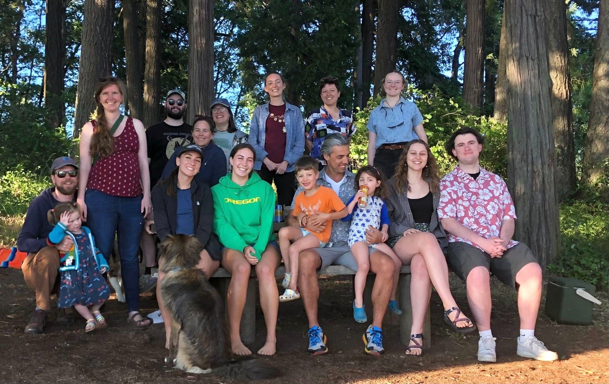 The lab group clusters around and on a picnic table set among tall trees. Everyone is smiling at the camera.