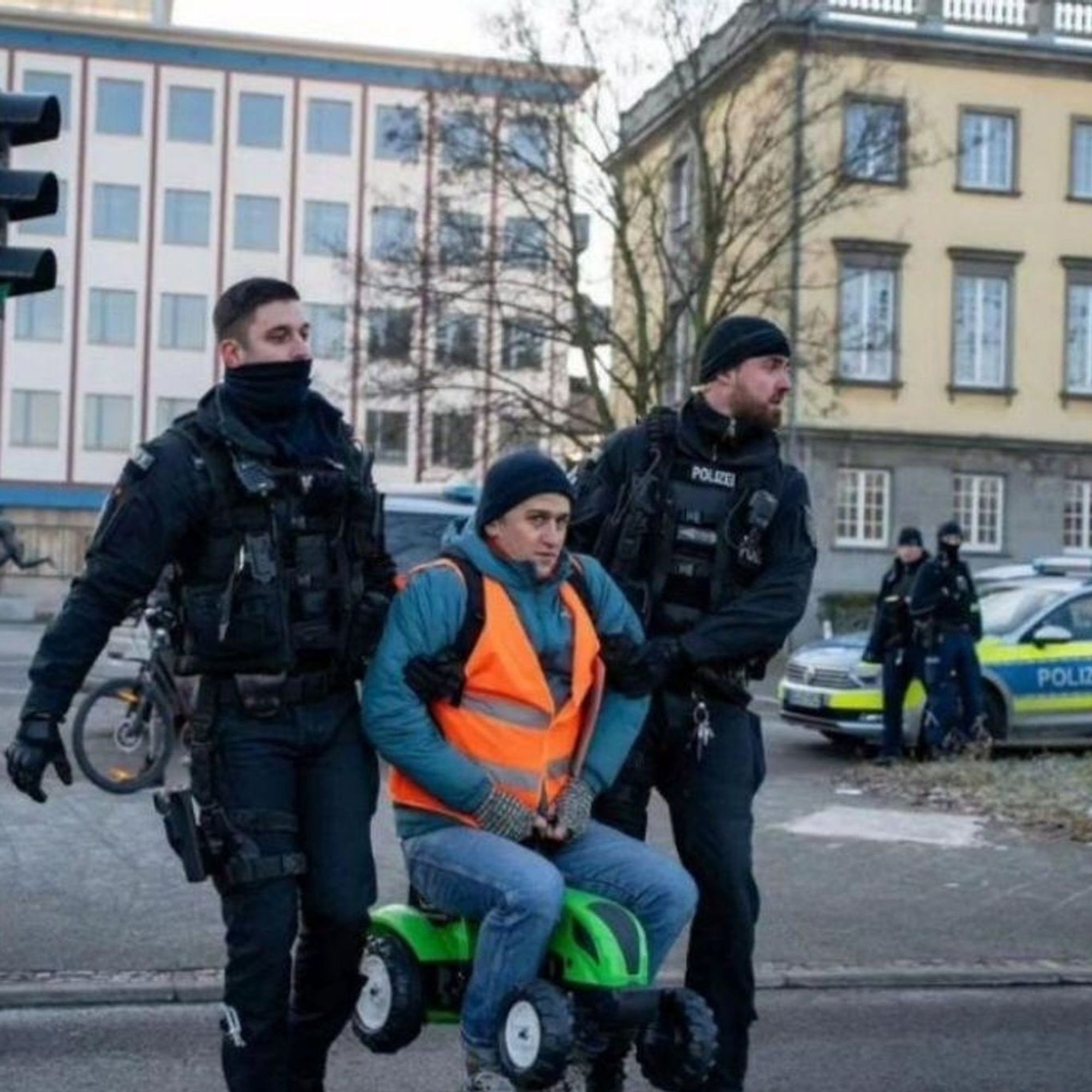 Eine Blockade-Demo der Letzten Generation: ein Aktivist auf einem Tret-Traktor wird von zwei Polizisten aus der Blockade geräumt. Im Hintergrund rechts steht ein Polizeiwagen.