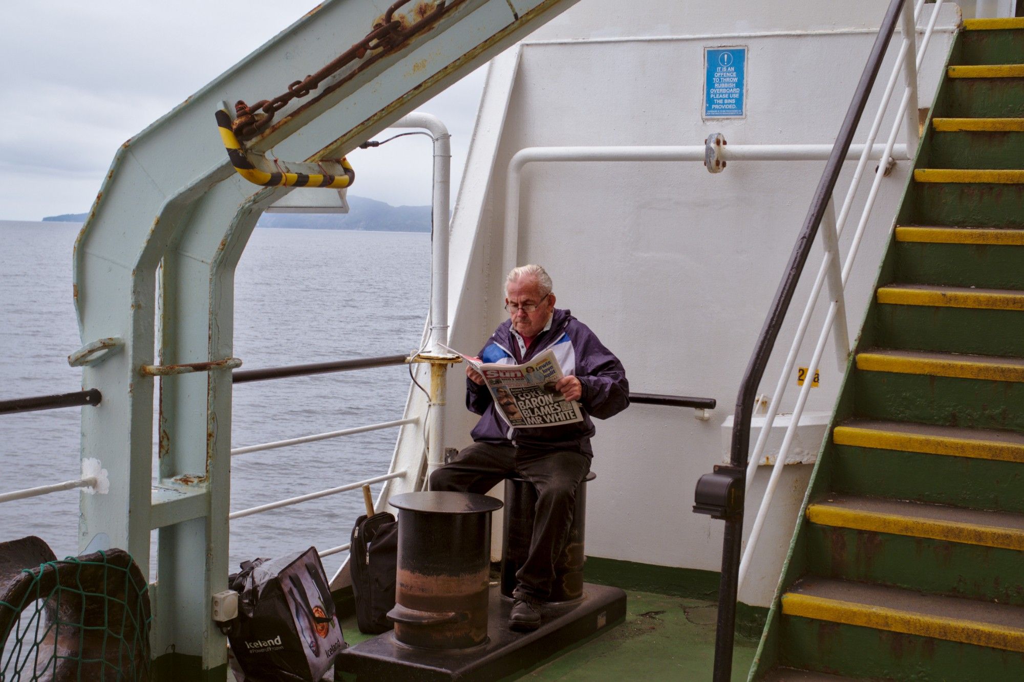 A man reading the Sun "newspaper" on the deck of a ferry. The grey sky overhead.