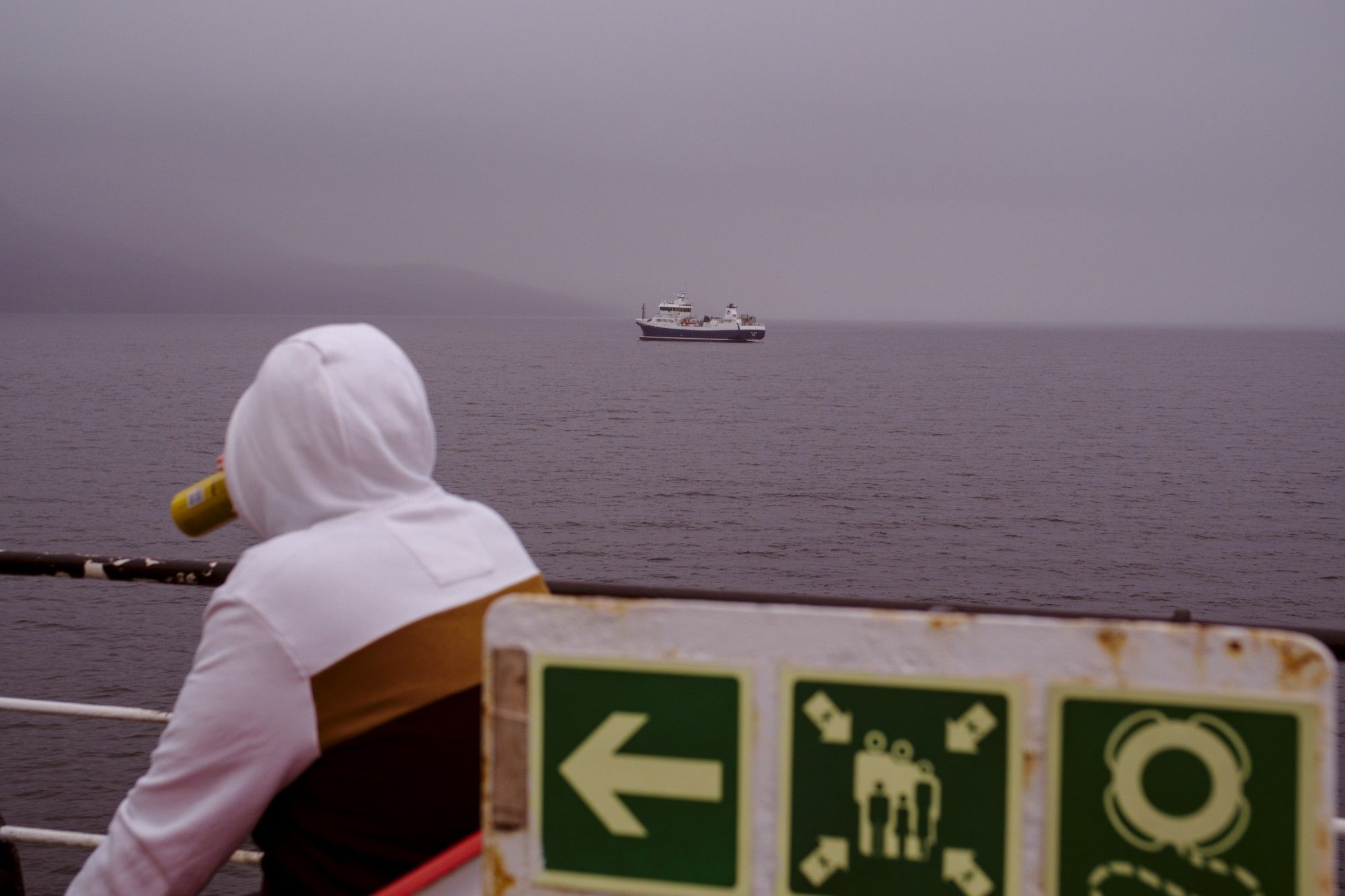 A man drinks from a can of beer onboard a ferry, an anchored fishing vessel is seen in the background. The sky is grey, the coast is covered by clouds.