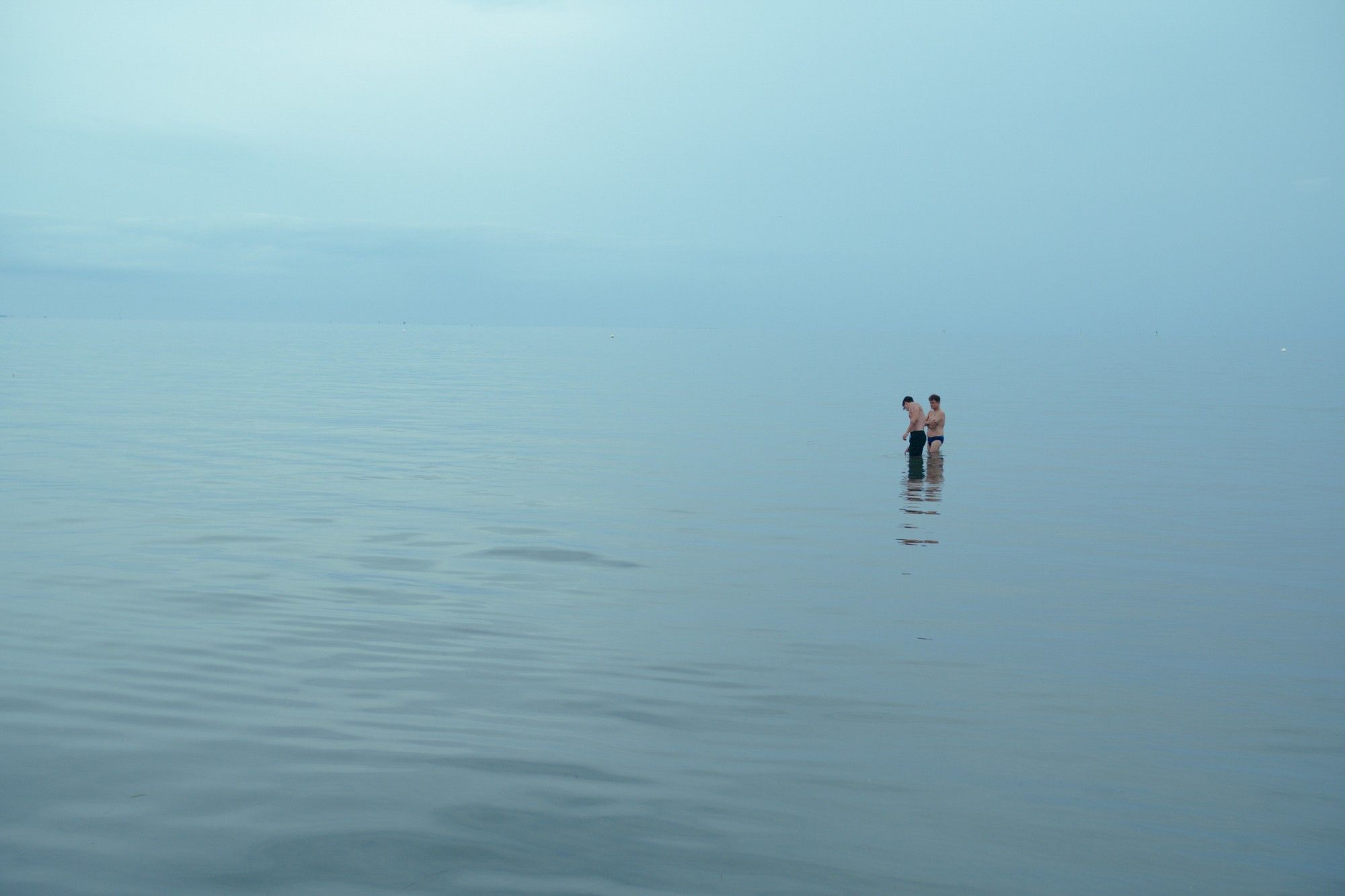 Photo of two men standing in shallow water. Nothing can be seen around them but the still ocean, the horizon melts into the misty sky.