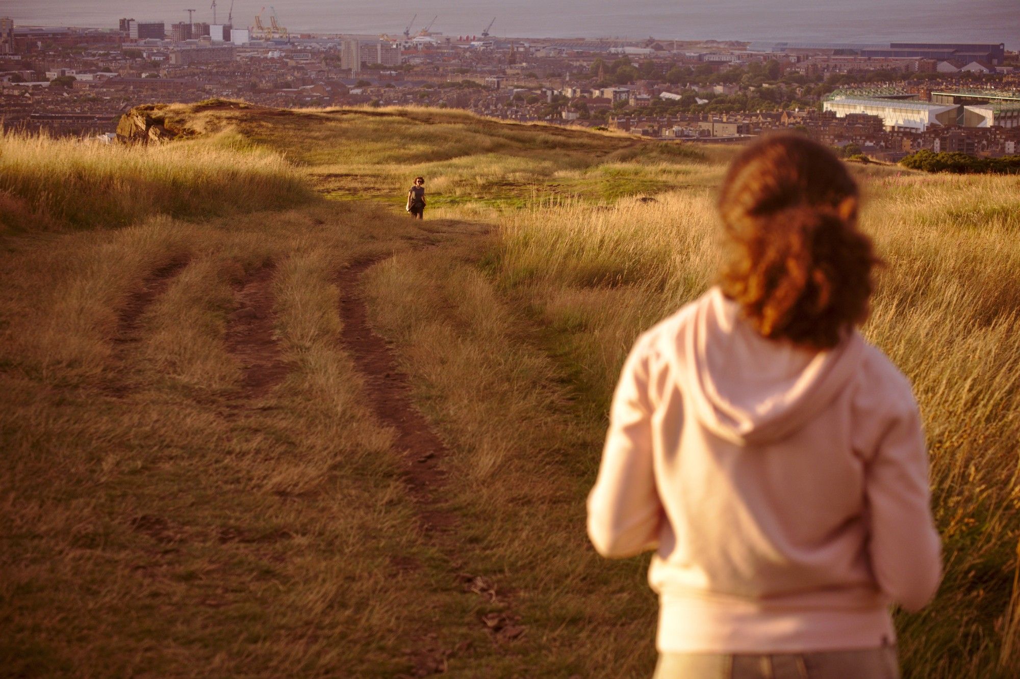 Evening shot of a hill in Holyrood Park overlooking Edinburgh. A girl, seen from behind, is facing a person in the distance, the surrounding city is visible in the background.