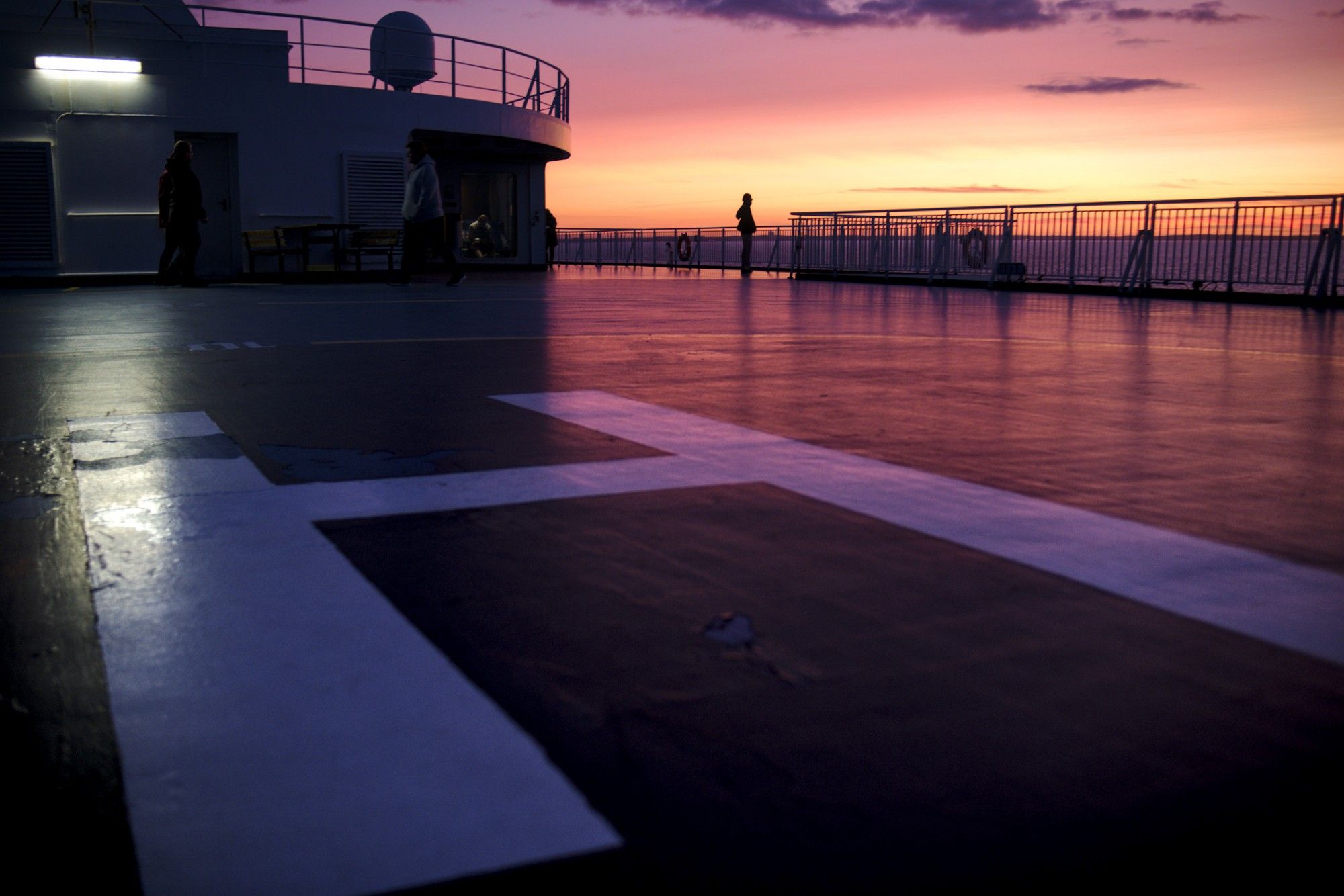 Silhouette of a person against a colourful sunset, on the floor in front, a large "H" indicating a helicopter landing area is visible.