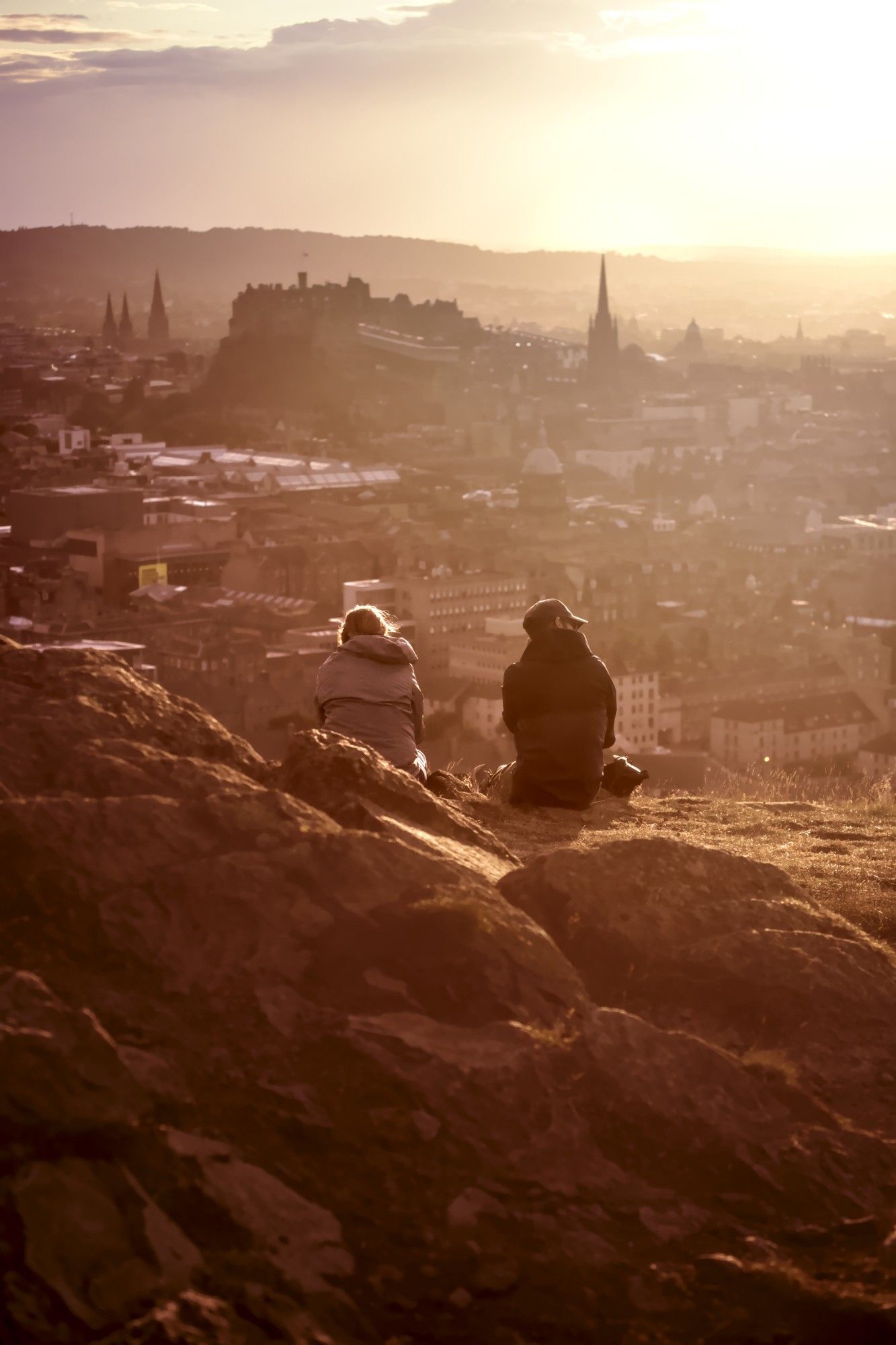 Evening shot of a hill in Holyrood Park overlooking Edinburgh. Two onlookers watch the sunset over the city.