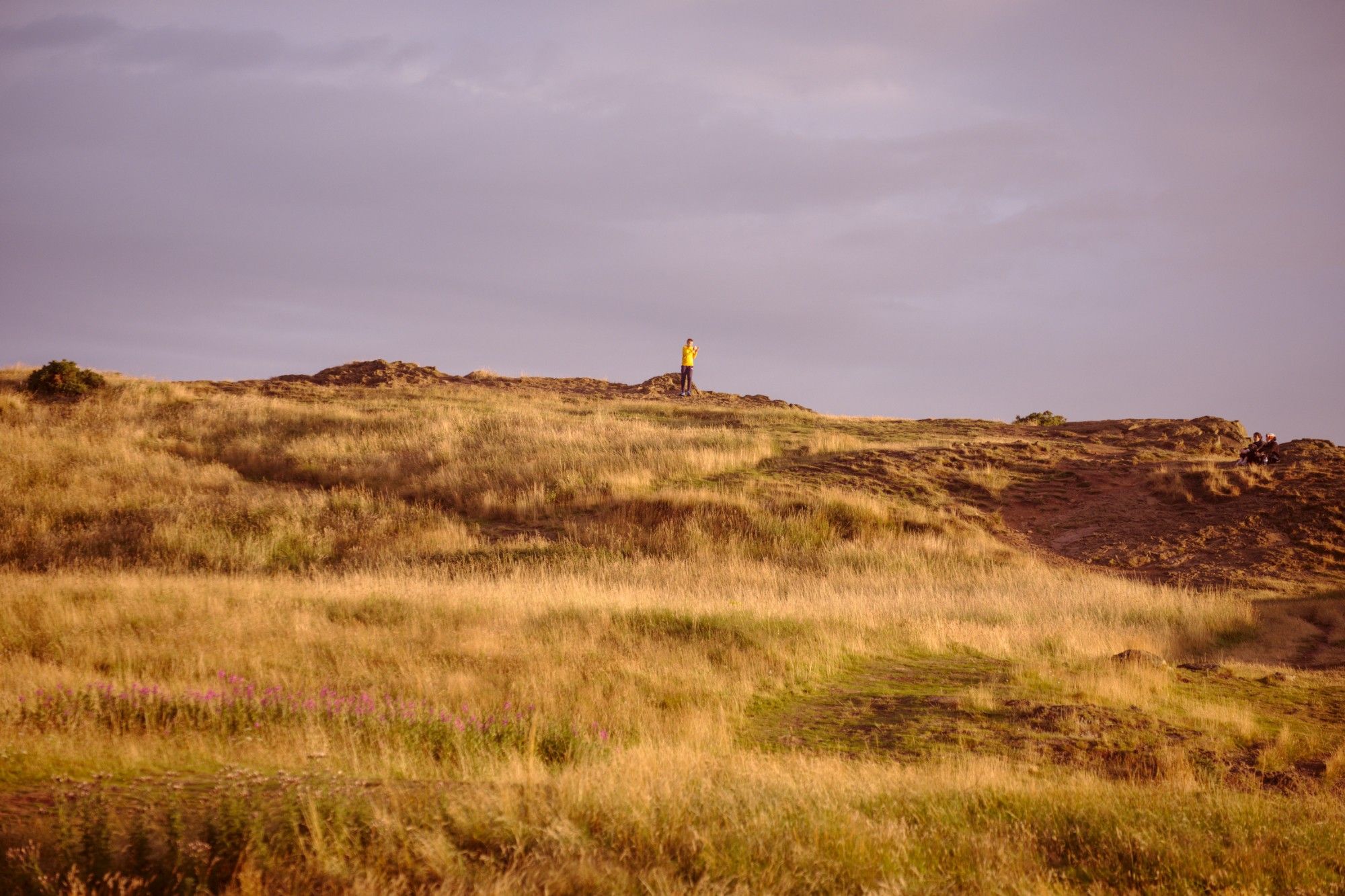 Evening shot taken on a hill in Holyrood Park, Edinburgh. A person in a yellow sweater on the grassy hill, illuminated by the warm evening sun, against a blue-grey sky.