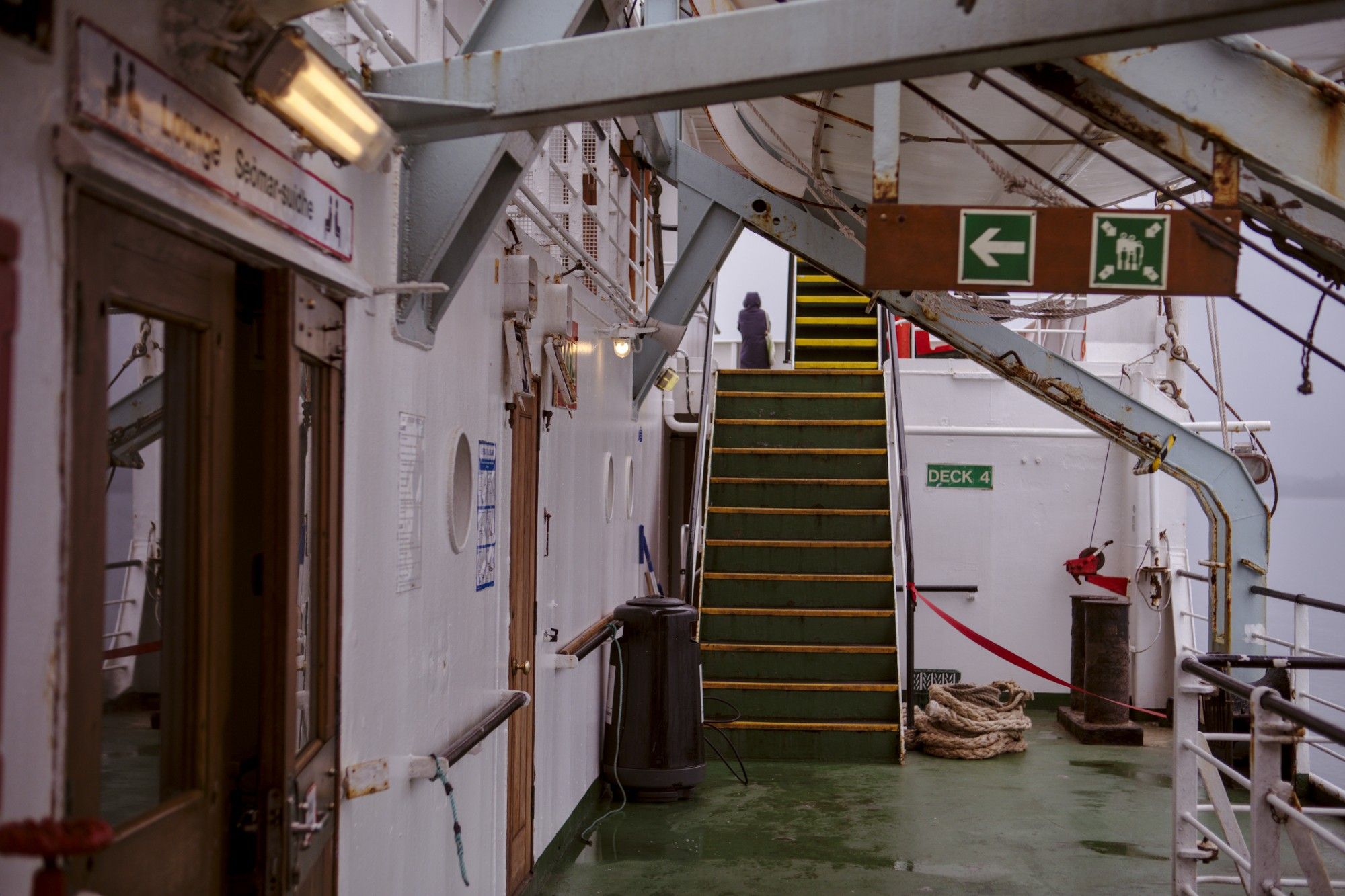 The deck of a ferry which has seen better days, wet from rainwater, a dark grey scene. In the background, a passenger is seen, a life boat can be seen at the top.