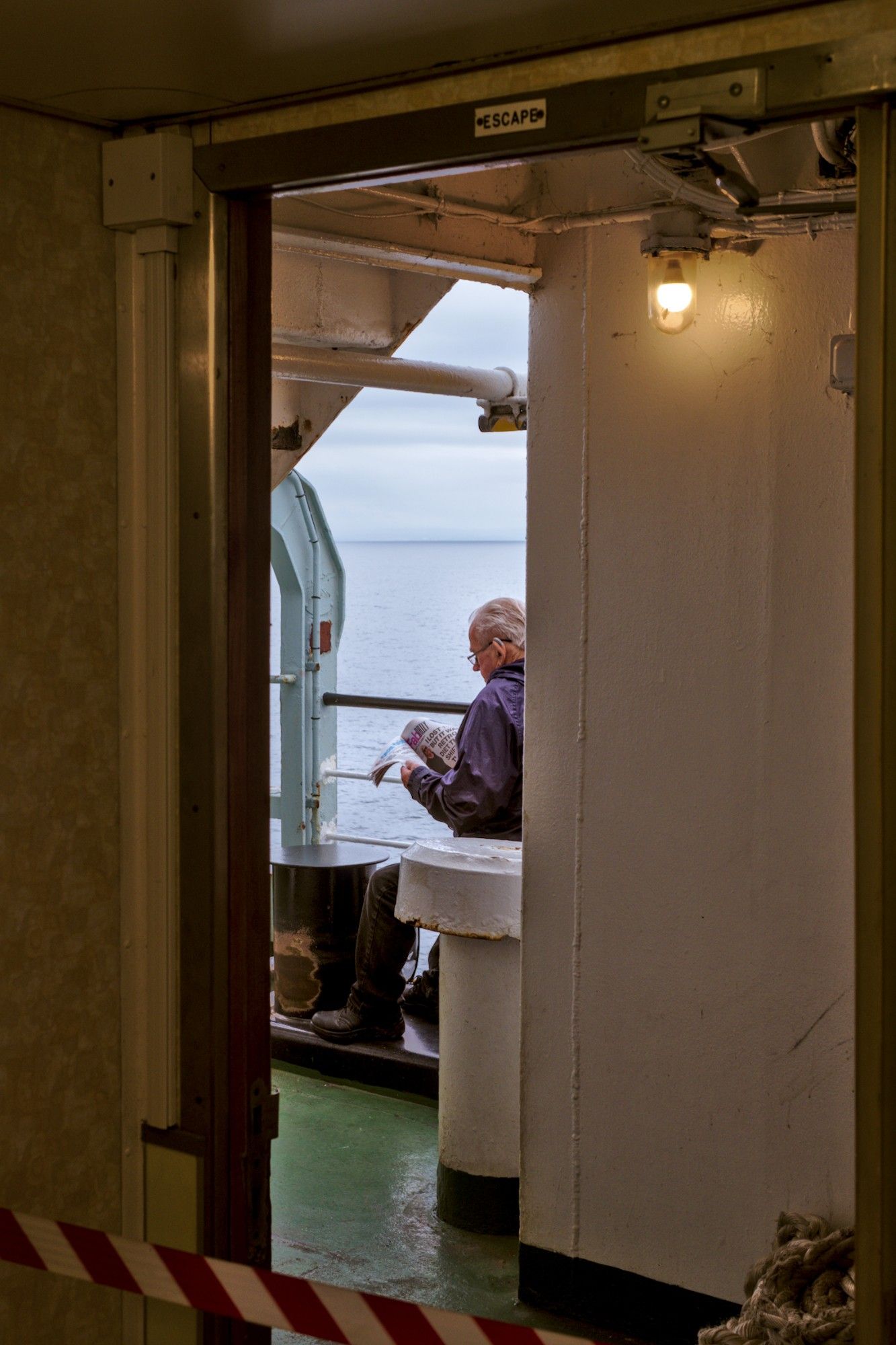 A man reading a newspaper onboard the ferry from Ardrossan to Brodick, Isle of Arran, Scotland. The man is seen through an open door of the ship, the sea is visible behind him.