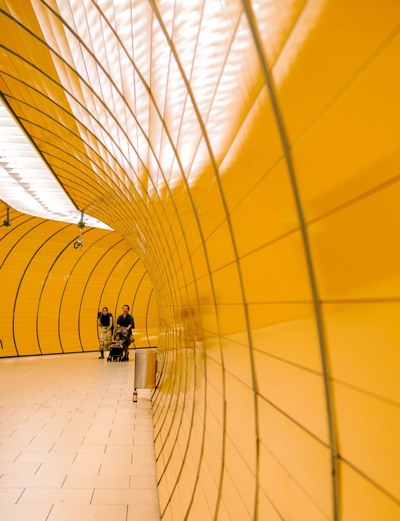 An orange-tiled metro station, a couple with a stroller are coming through the round passenger tunnel.