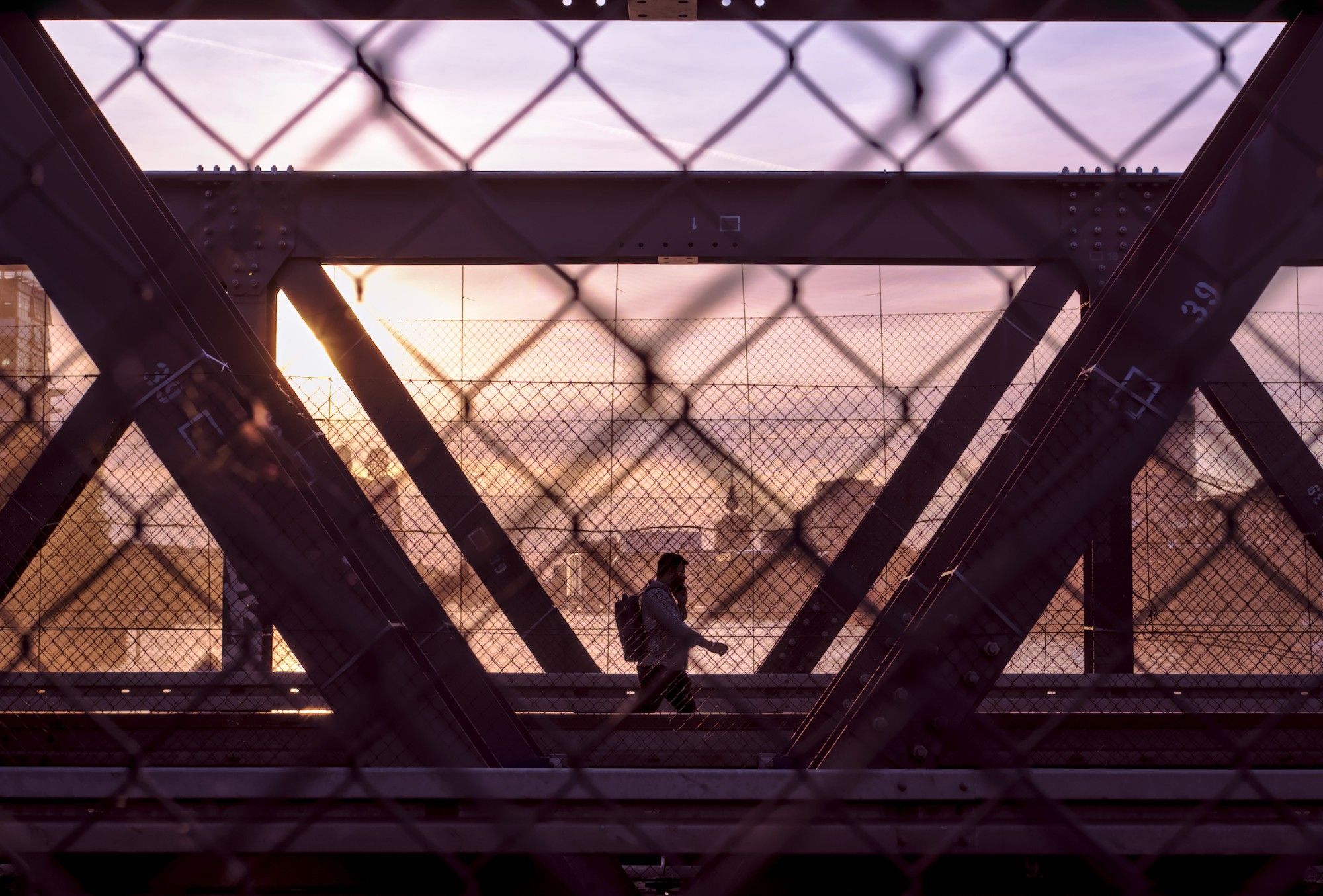 A man walking over a bridge below an evening sky; steel beams and mesh wire in the foreground and overhead, the silhouette of the city in the background. Taken on Elsenbrücke in Berlin.