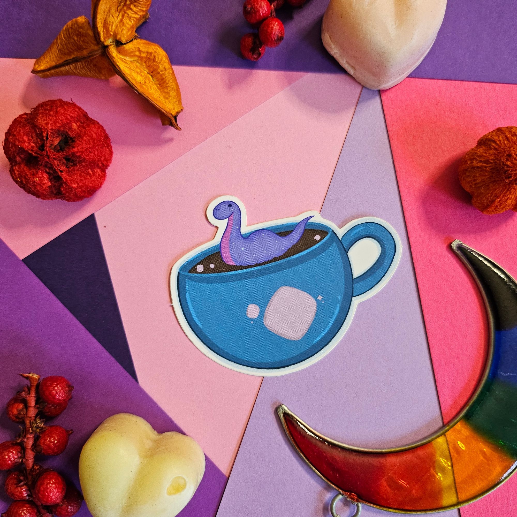 a photo of a sticker that is a small loch ness creature in a large cup of hot chocolate. the cup is blue with a marshmallow on it. there is a colouful back ground with some cute clutter around the sticker.