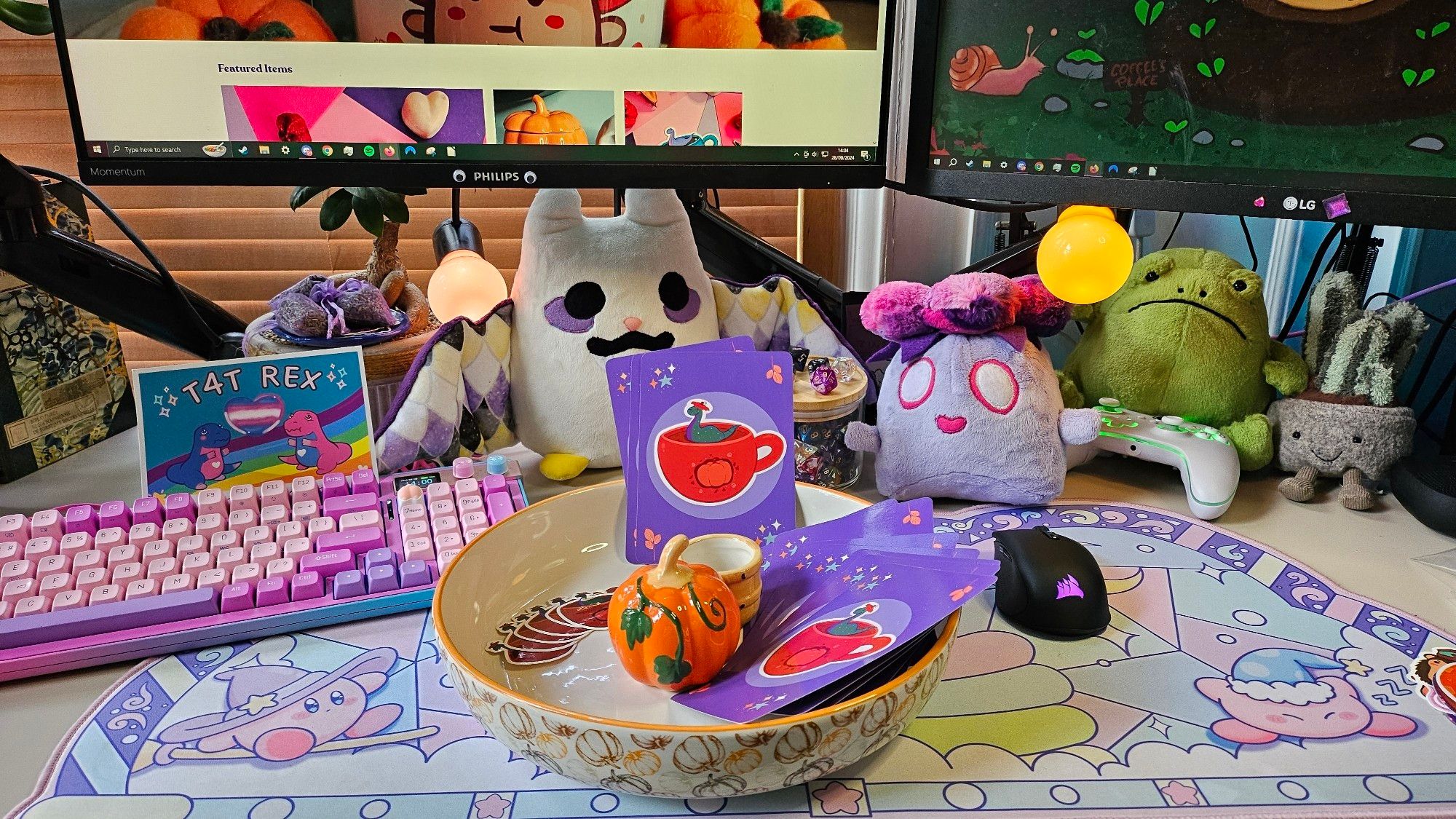 A postcard of Nessie swimming on an orange tea cup with an autumn pumpkin motiv on its front. Nessie is wearing the top of a pumpkin as a hat (like the portion you cut out around the stalk when you make a Halloween lantern). The postcard has a purple background and some floral ornaments in the corners. The cards are arranged in an autumn themed serving bowl with a ceramic sculpted pumpkin in its middle on my colourfully decorated work desk full of plushies, a pink and teal keyboard and a Kirby desk mat.