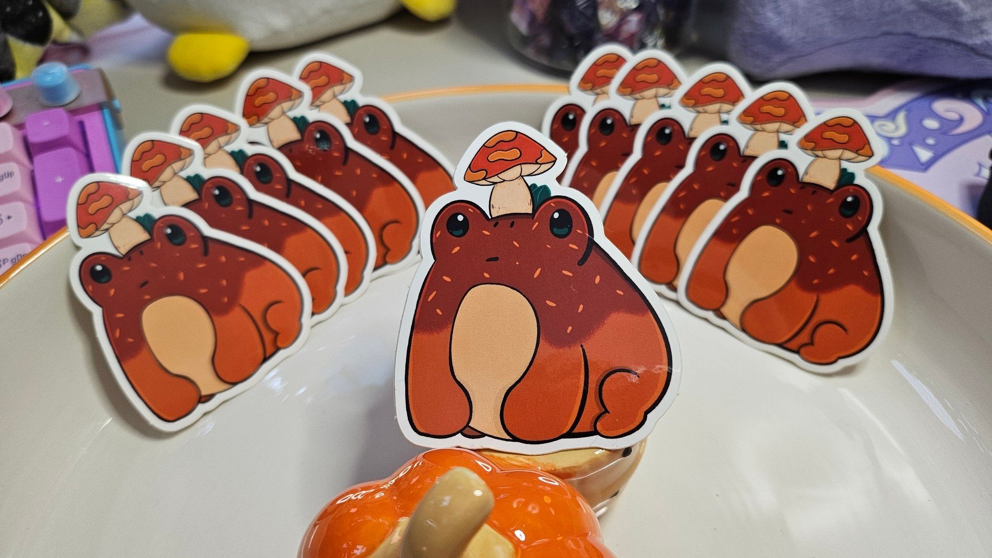 A die-cut sticker of a chubby cartoon frog with a facial expression that seems to say "I'm not sure about this". xD
The frog is pumpkin orange on the bottom of their legs and but, a darker orange with lighter sprinkles on top of their body and face. They have a red cap mushroom on their head like a hat.