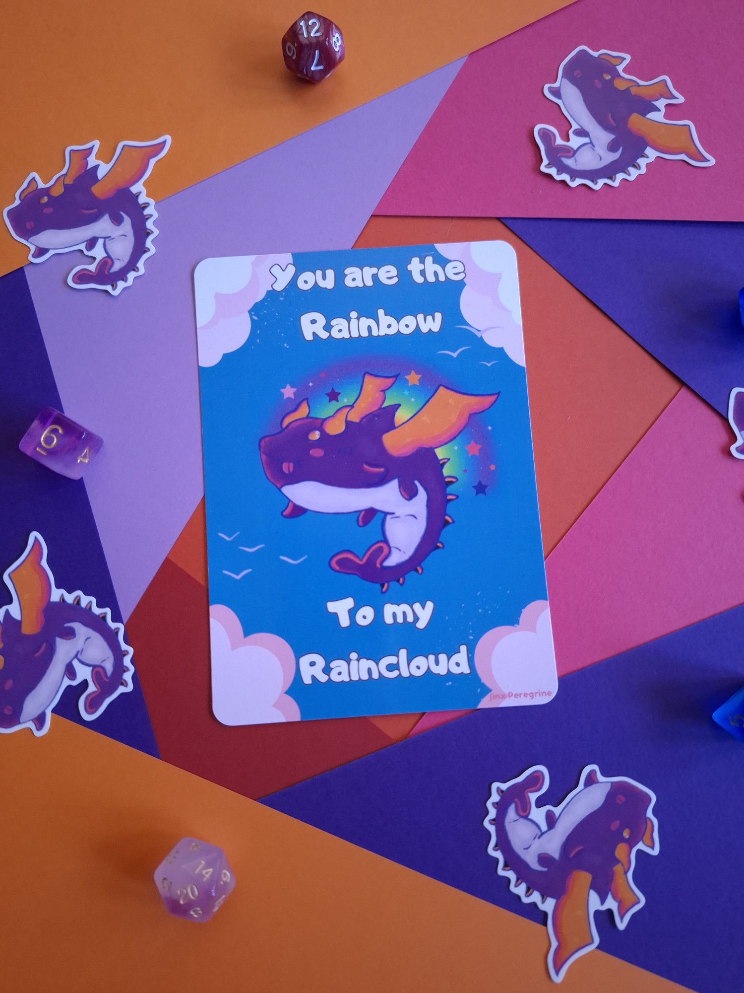 a photo of a postcard with a fantastical creature called a shragon, part shark, part dragon,they are purple with yellow wings, yellow horns, that is flying in a blue sky with clouds around them. The words "you are the rainbow to my raincloud is around them in a white bubble font. There are stickers of the same shragon around the postcard on a colourful table.