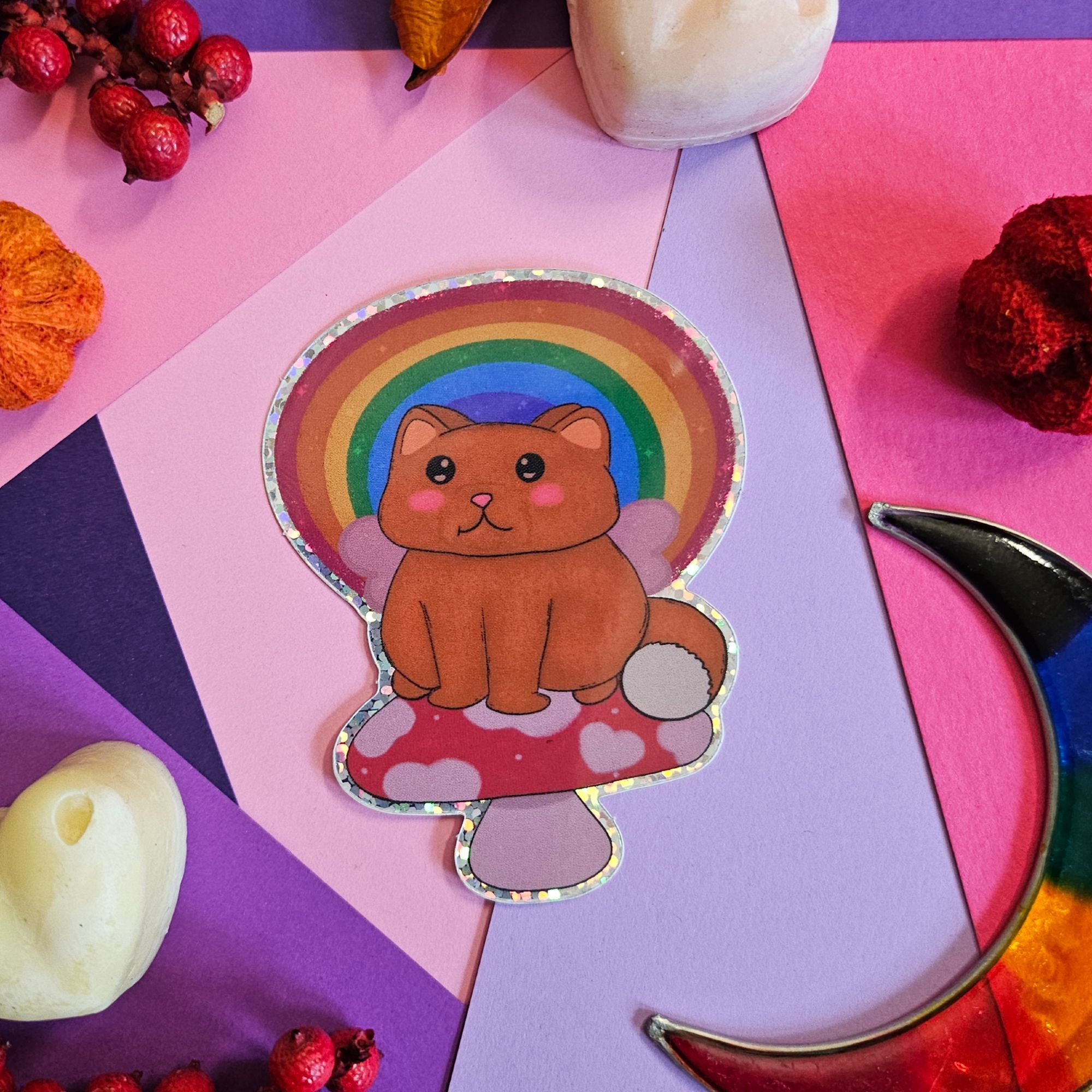 a photo of a sparklie sticker of a orange cat with pink fairy wings sitting on red cap mushroom with rainbow behind them.