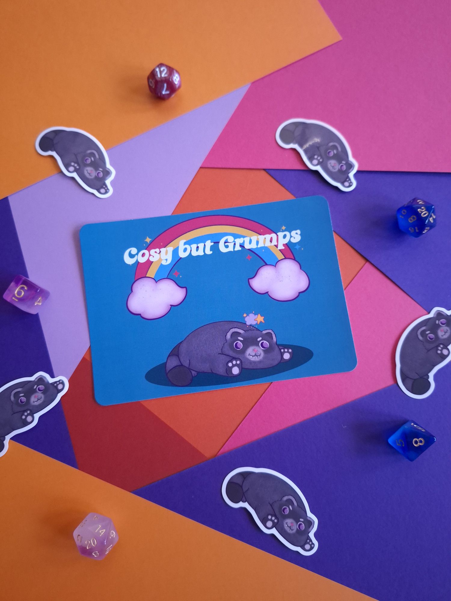 a photo of a postcard that has a blue back ground and cute art of a raccoon cat. the words "cosy but grumps" is over their head in front of a rainbow. there are racoon cat stickers around the postcard on a colouful table.