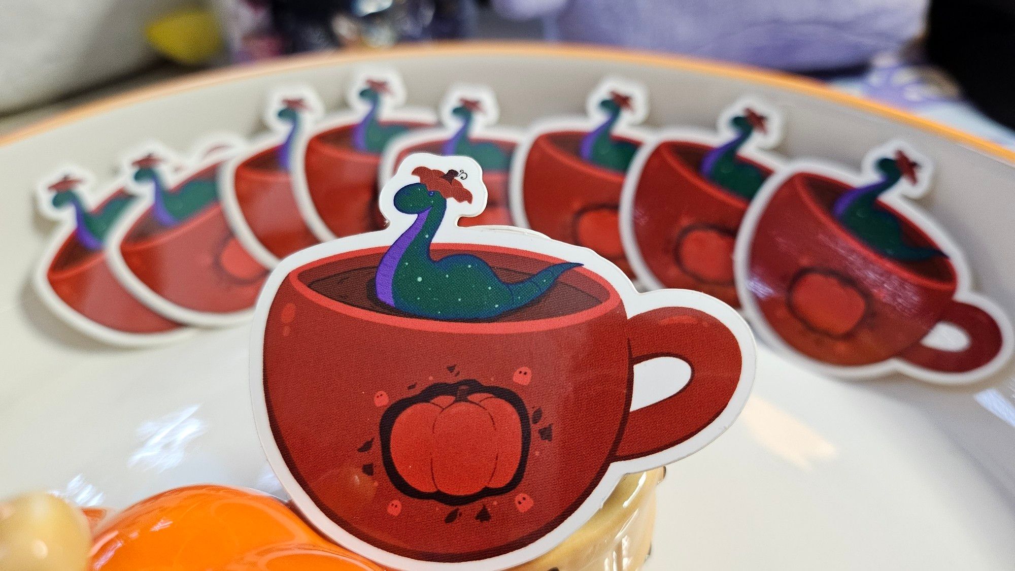 A die-cut sticker of Nessie swimming on an orange tea cup with an autumn pumpkin motiv on its front. Nessie is wearing the top of a pumpkin as a hat (like the portion you cut out around the stalk when you make a Halloween lantern).