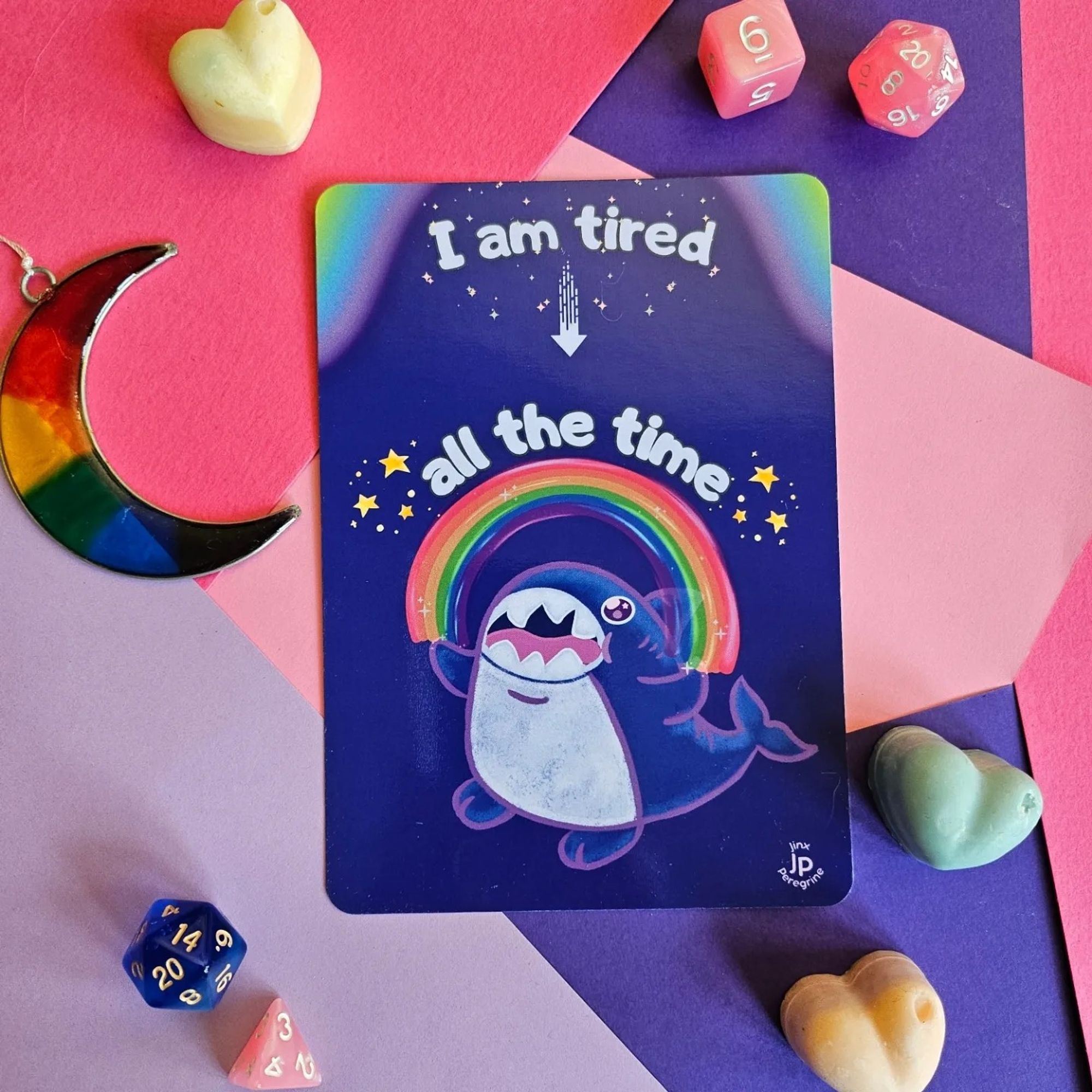 A postcard with a cute and grumpy shark friend with an open mouth as though they're shouting, who is spanning a rainbow across their outstretched fins. There is a caption above them and the rainbow, which reads: "I am tired all the time"