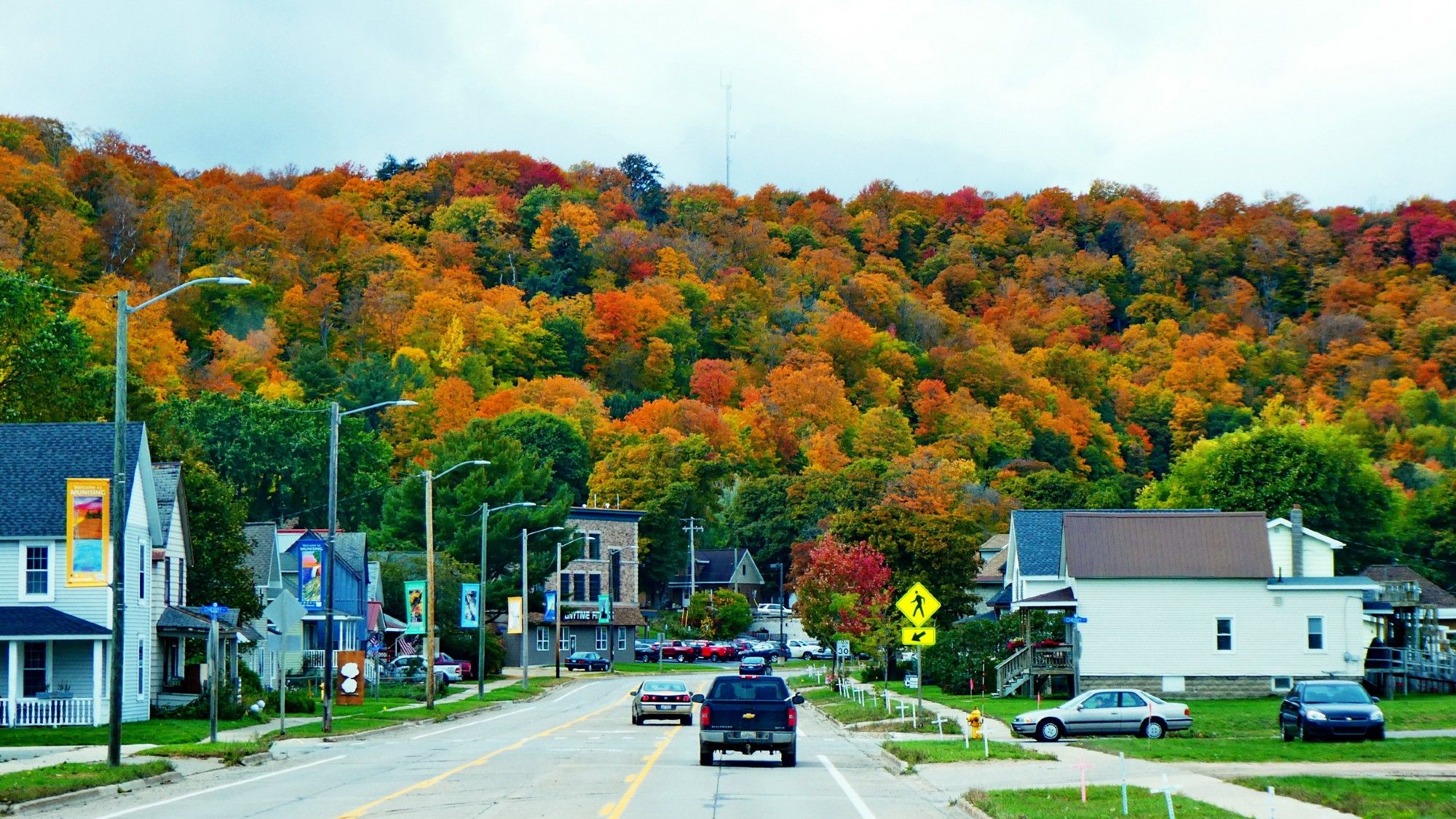 Une route dans le village de Munising. Quelques maisons de chaque côté. Au bout, la route vire fortement vers la droite et dans l'extérieur du tournant, Une grande colline où une forê dense laisse son feuillage éclater ses couleurs d'automne. Vert, jaune, brun, orangé et quelques traces de rouge sont à l'honneur.