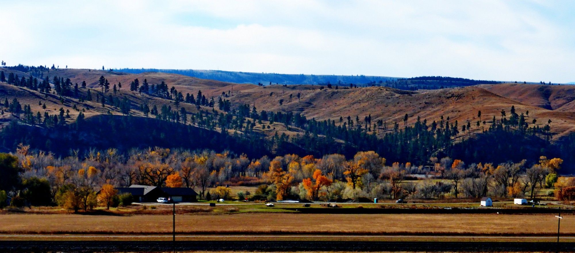 Un panorama de terres agricoles vidées de leur récolte et prêtes à être retravaillées au printemps, En toile de fond, des collines parsemées d'arbres et entre les deux, une ferme et des arbres, Certains sont gris car ayant déjà perdu tout leur feuillage alors que d'autres sont encore flamboyant de leurs couleurs d'automne. le contraste est saisissant.