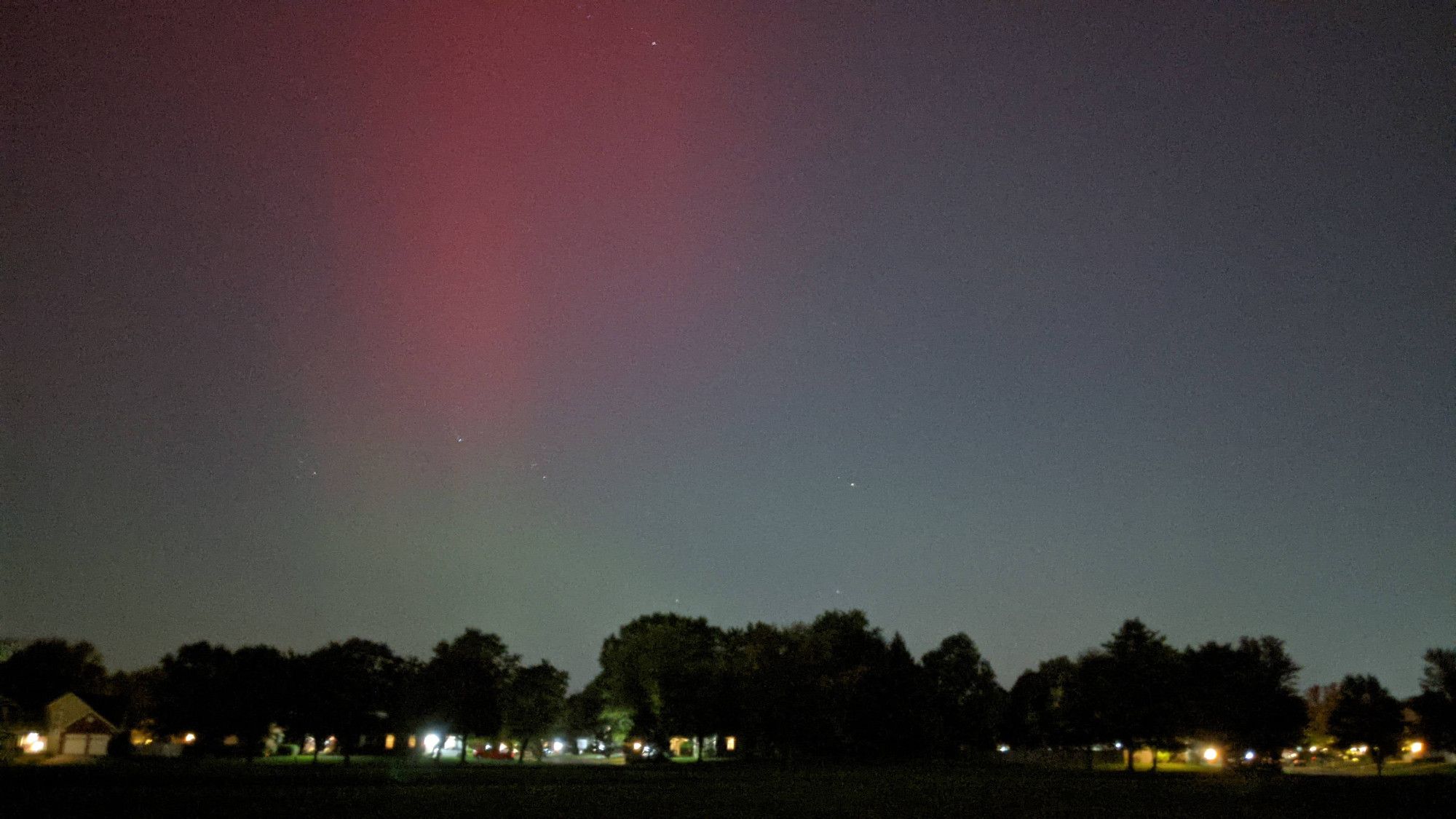 Picture of the northern lights in a dark sky. Orange/red cloud in the sky fading into blue green at the horizon with dark trees and the lights of houses at the bottom of the frame.