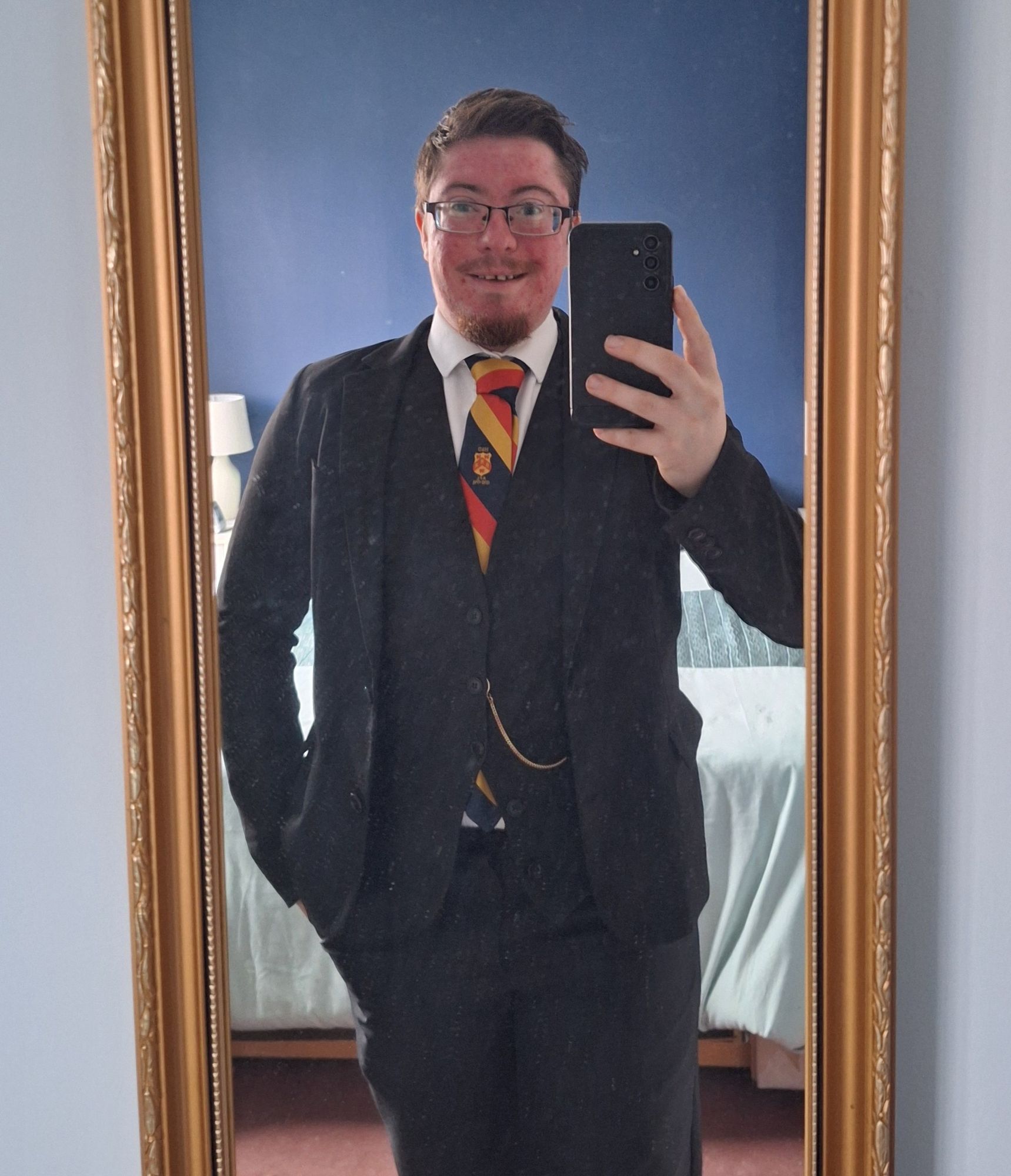 Nicholas wearing a three piece suit,  with a white shirt, a black, yellow and red striped tie and the chain of a pocketwatch visible. He is smiling in sheer joy at his reflection