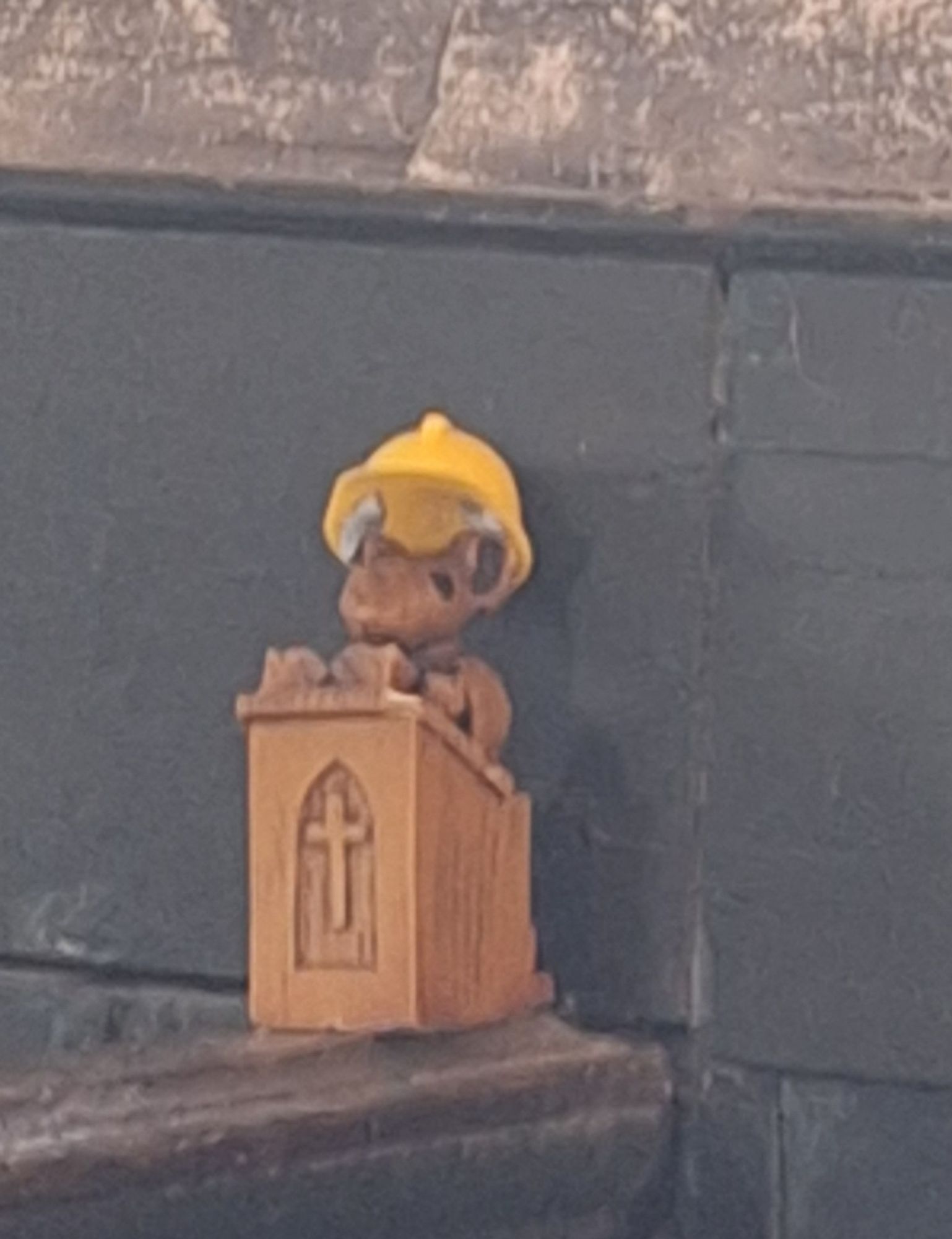 A small carved wooden mouse behind a wooden pulpit wearing a tiny yellow hard hat