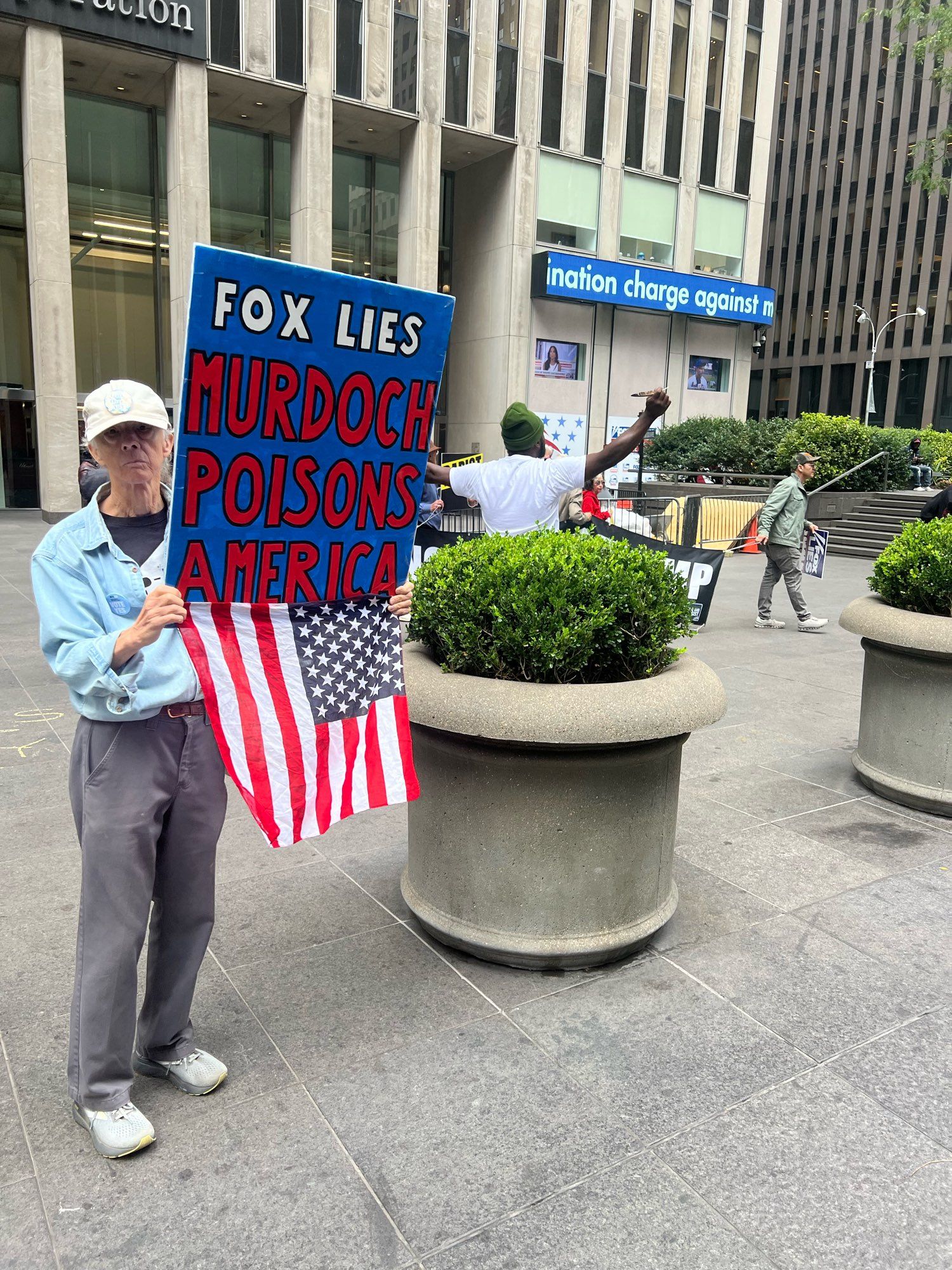 A person protesting in front of Fox News Corp. with an American flag and a sign that says, “Fox lies, Murdoch poisons America”