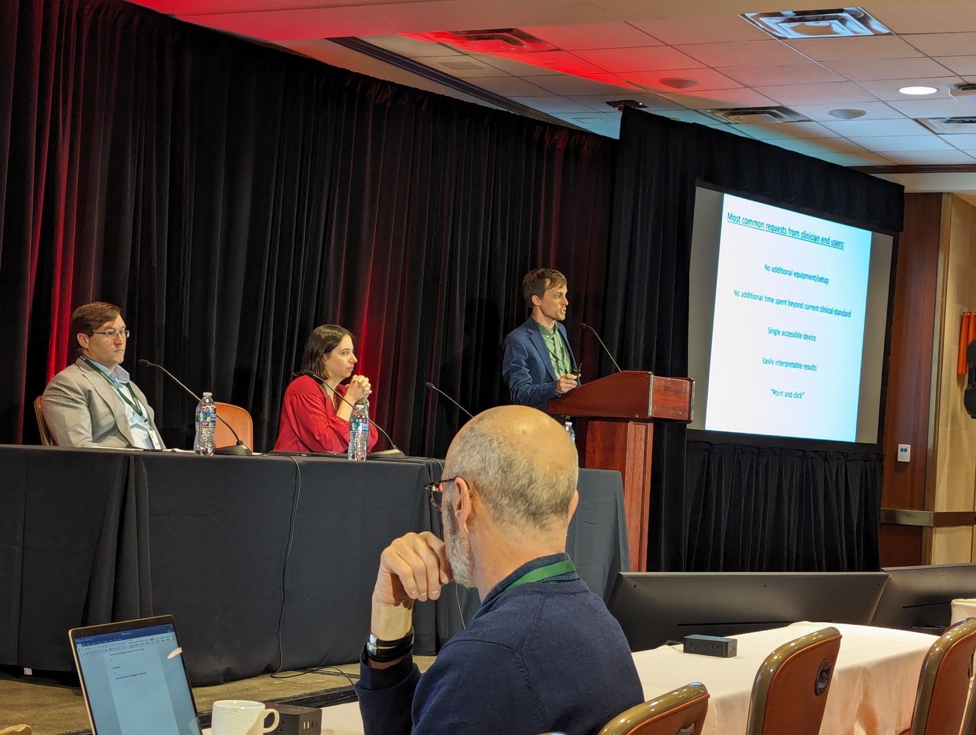 Ryan is standing at a lectern onstage looking out towards the audience. The slides are displayed on the right, and the other two presenters in the session are seated at a table onstage to the left.