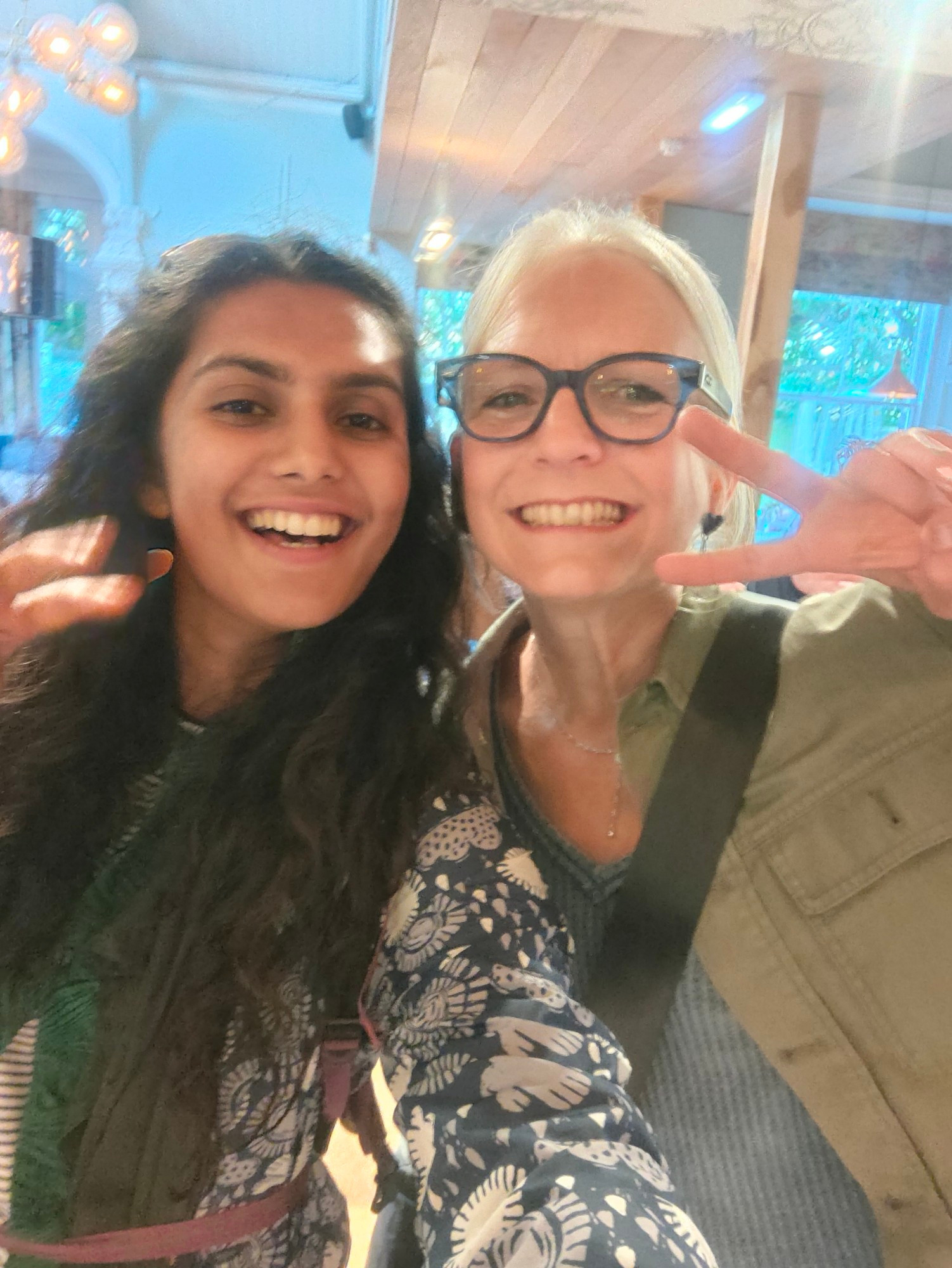 Photo of Asian woman with long black hair and blonde woman with framed glasses holding the peace signs to camera