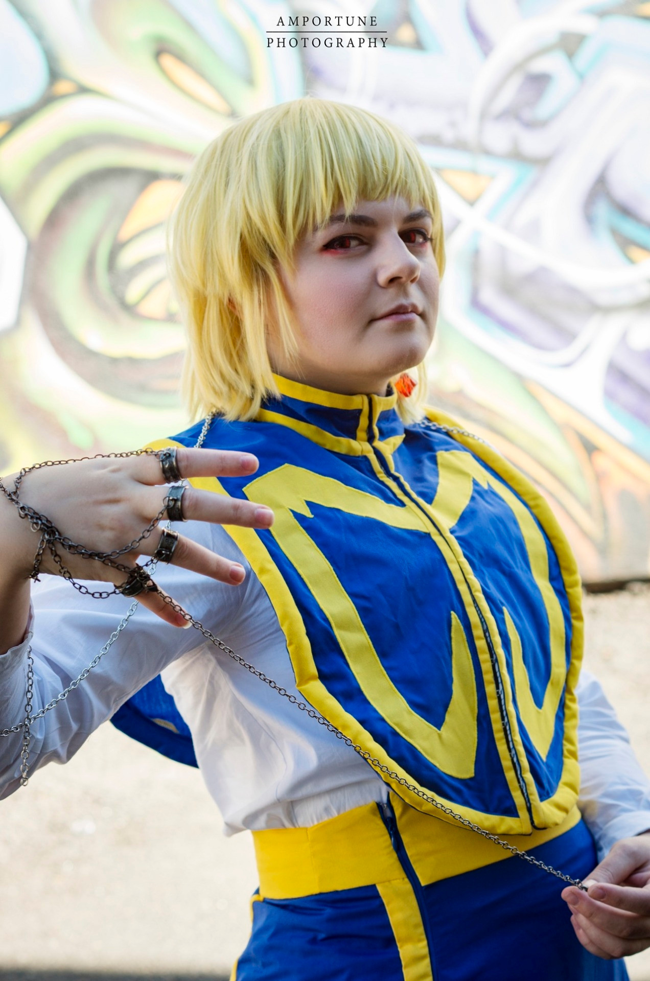 Cosplayer dressed as Kurapika from Hunter x Hunter; she's wearing a long blue skirt with yellow trim, a white undershirt, and a blue and yellow top. It is a waist up image in front of a graffiti wall, and she is holding a chain accessory.