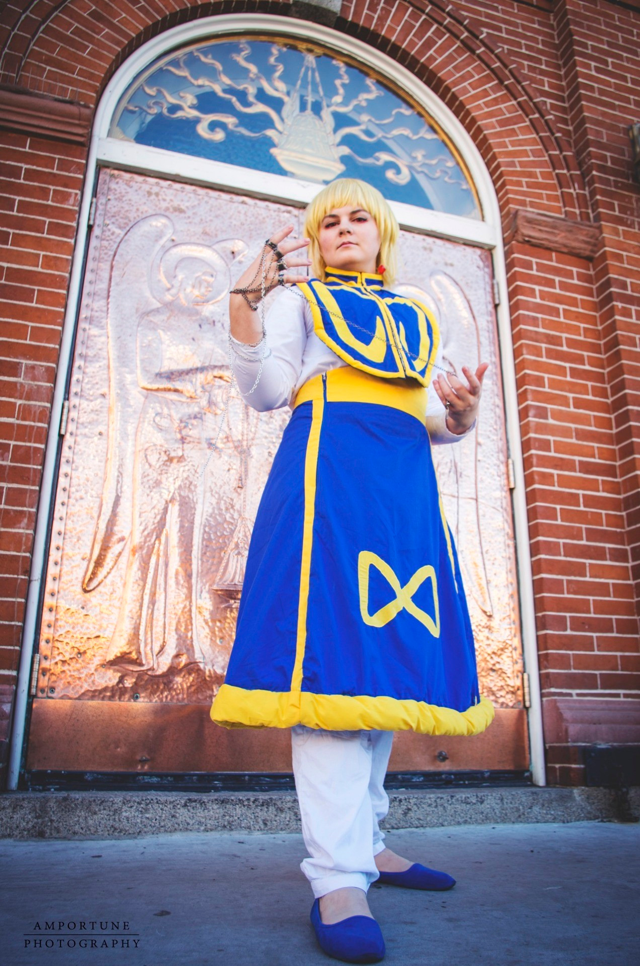 Cosplayer dressed as Kurapika from Hunter x Hunter; she's wearing a long blue skirt with yellow trim and a yellow infinity design on the skirt, white pants, a white undershirt, and a blue and yellow top. She's standing at an angle looking down at the camera, in front of copper plated church doors, and is holding a chain accessory.