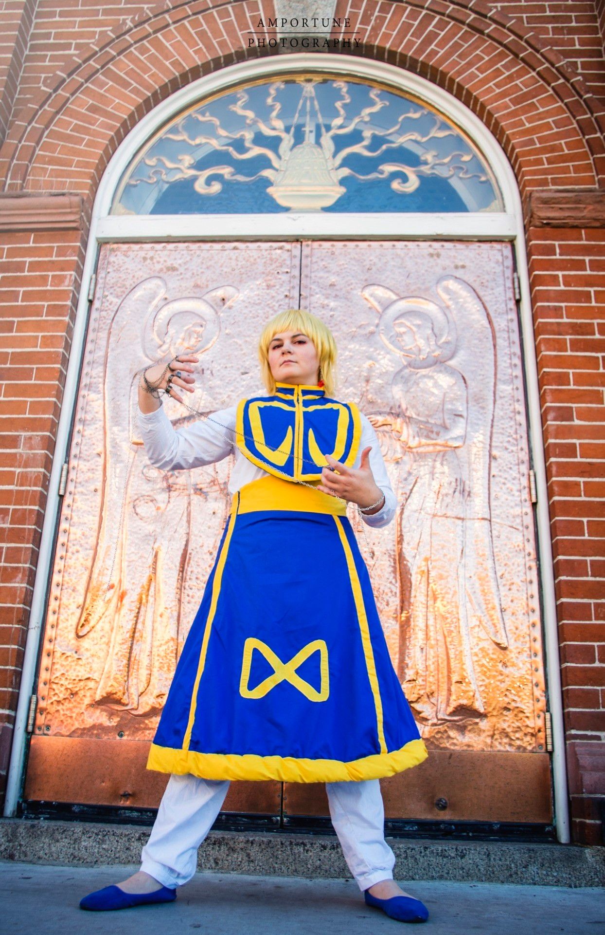 Cosplayer dressed as Kurapika from Hunter x Hunter; she's wearing a long blue skirt with yellow trim and a yellow infinity design on the skirt, white pants, a white undershirt, and a blue and yellow top. She's standing at an angle looking down at the camera, in front of copper plated church doors, and is holding a chain accessory.