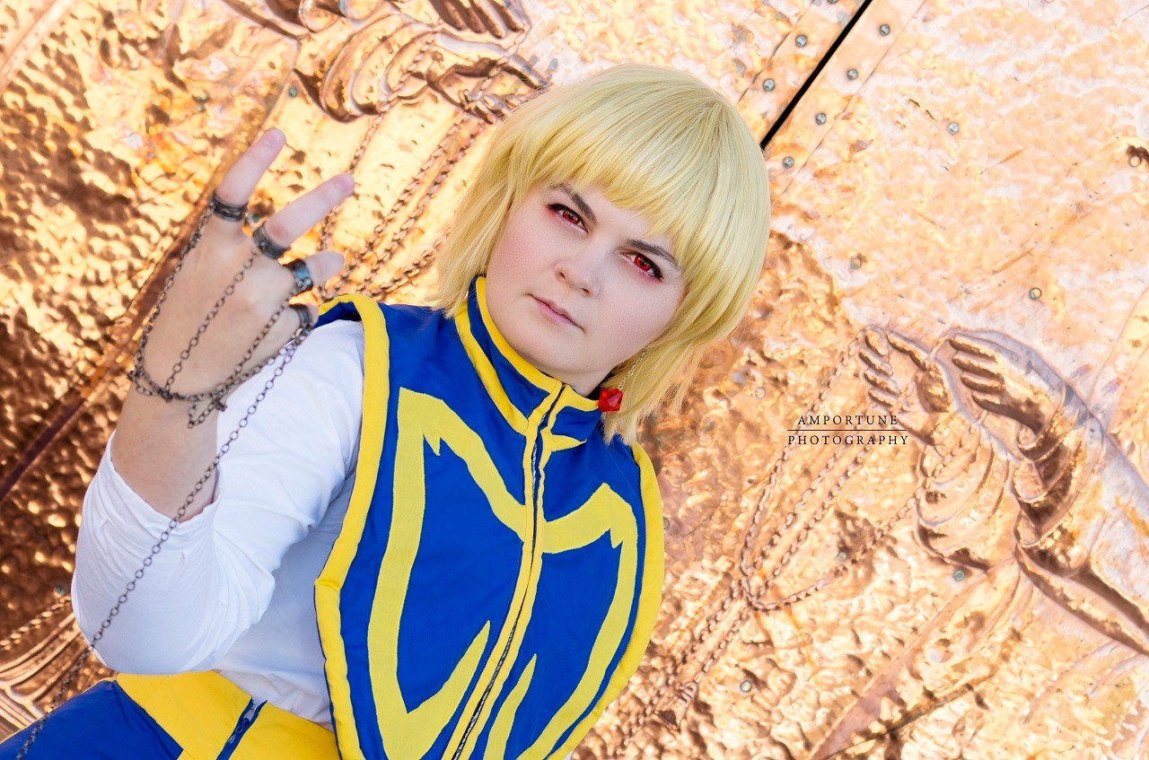 Cosplayer dressed as Kurapika from Hunter x Hunter; she's wearing a long blue skirt with yellow trim, a white undershirt, and a blue and yellow top. The photo is taken at an angle in front of copper plated church doors, and is holding a chain accessory.