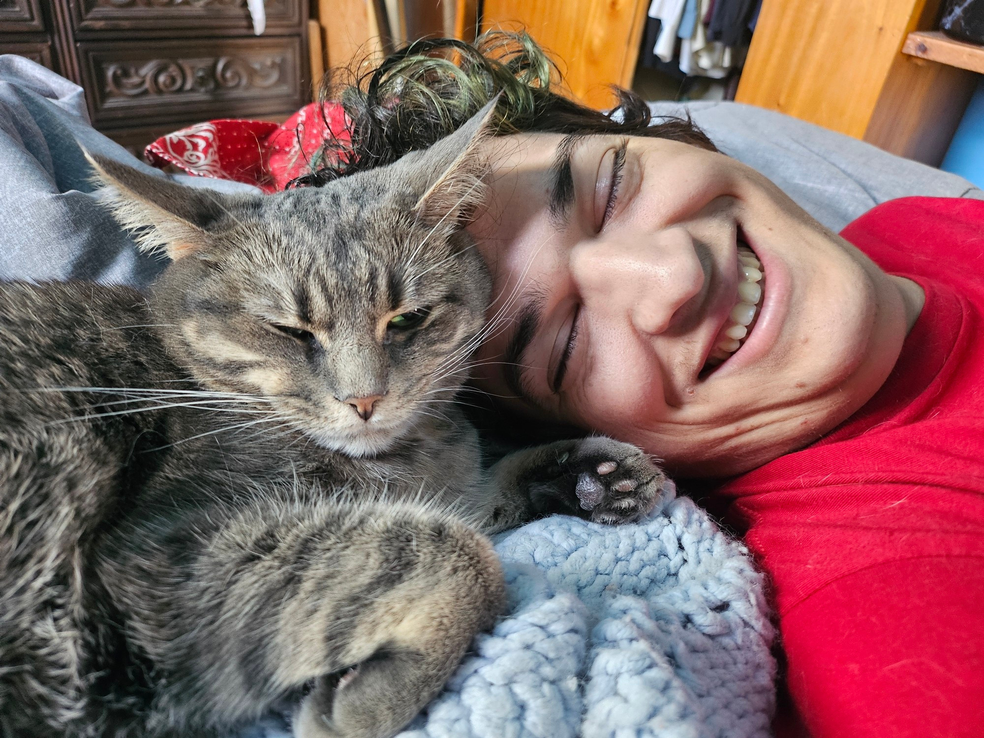 A photo of my gray tabby cat, Suki, laying up against the face of my partner, Art the TryHard, on a blue blanket on our bed