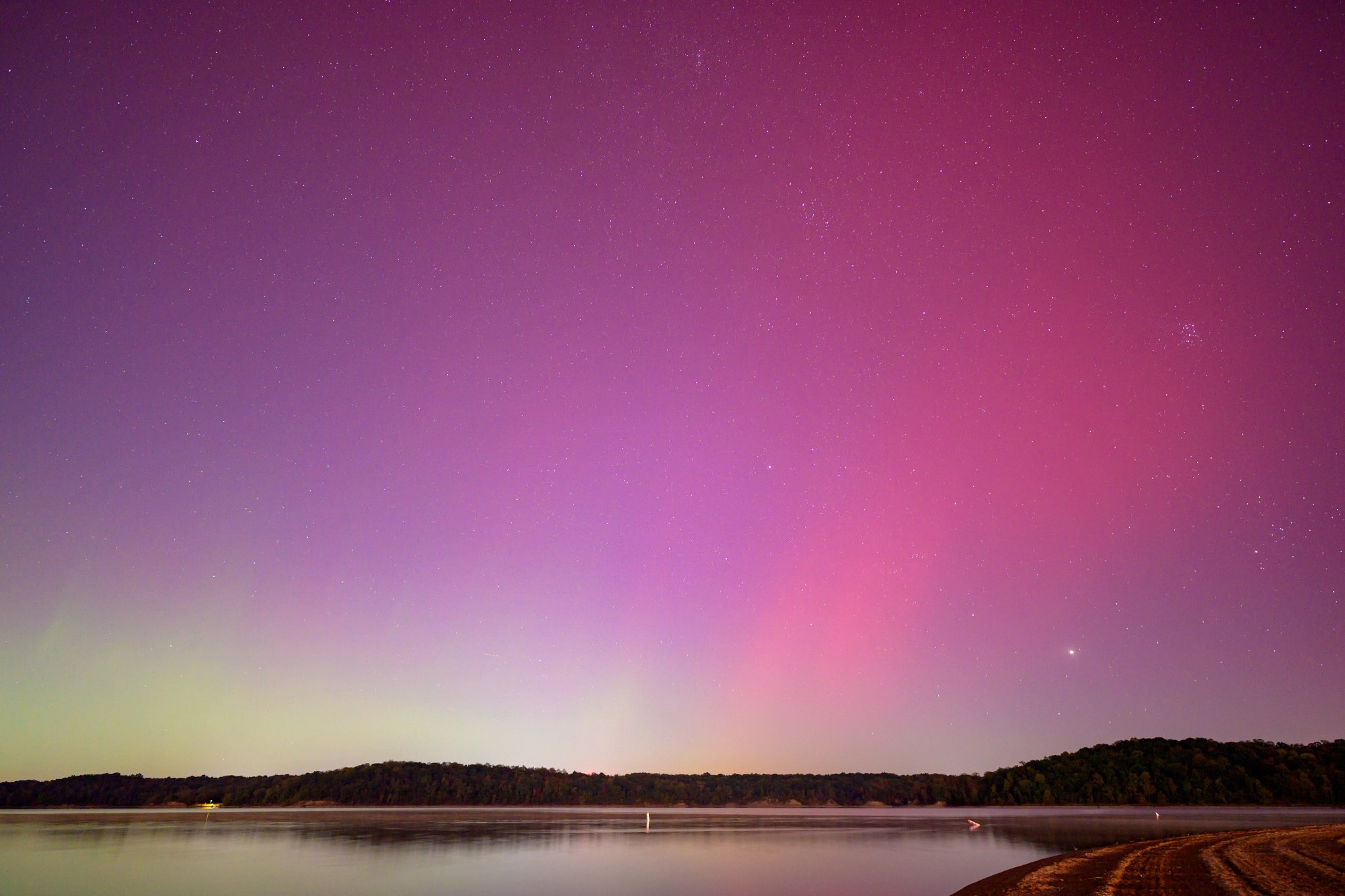 Aurora borealis as seen looking northeast over East Fork Lake at East Fork Lake State Park in Clermont County, Ohio (about 20 miles east-southeast of Cincinnati), on Thursday, October 10, 2024. A calm lake is in the foreground and a combed beach is at bottom right. The horizon on the opposite shore stretches low across the bottom of the photograph. The sky shows subtle green-yellow at left, pink at right, and purple above. Faint vertical streeks are visible throughout. What appears to be the largest visible star (near the bottom right of the sky) is actually the planet Jupiter. The Pleiades asterism is above and to the right of it.