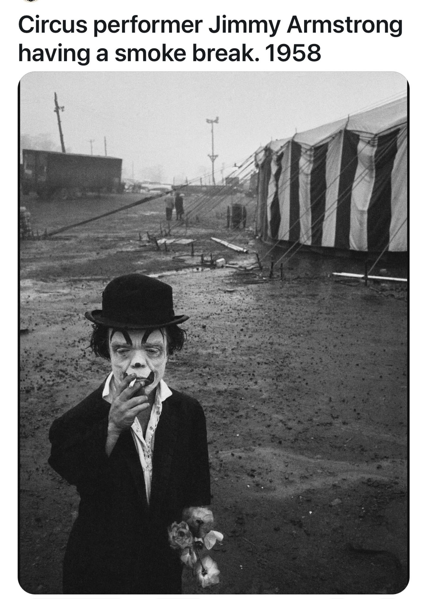 Photograph of a circus performer, standing alone outside the circus tent, smoking a cigarette