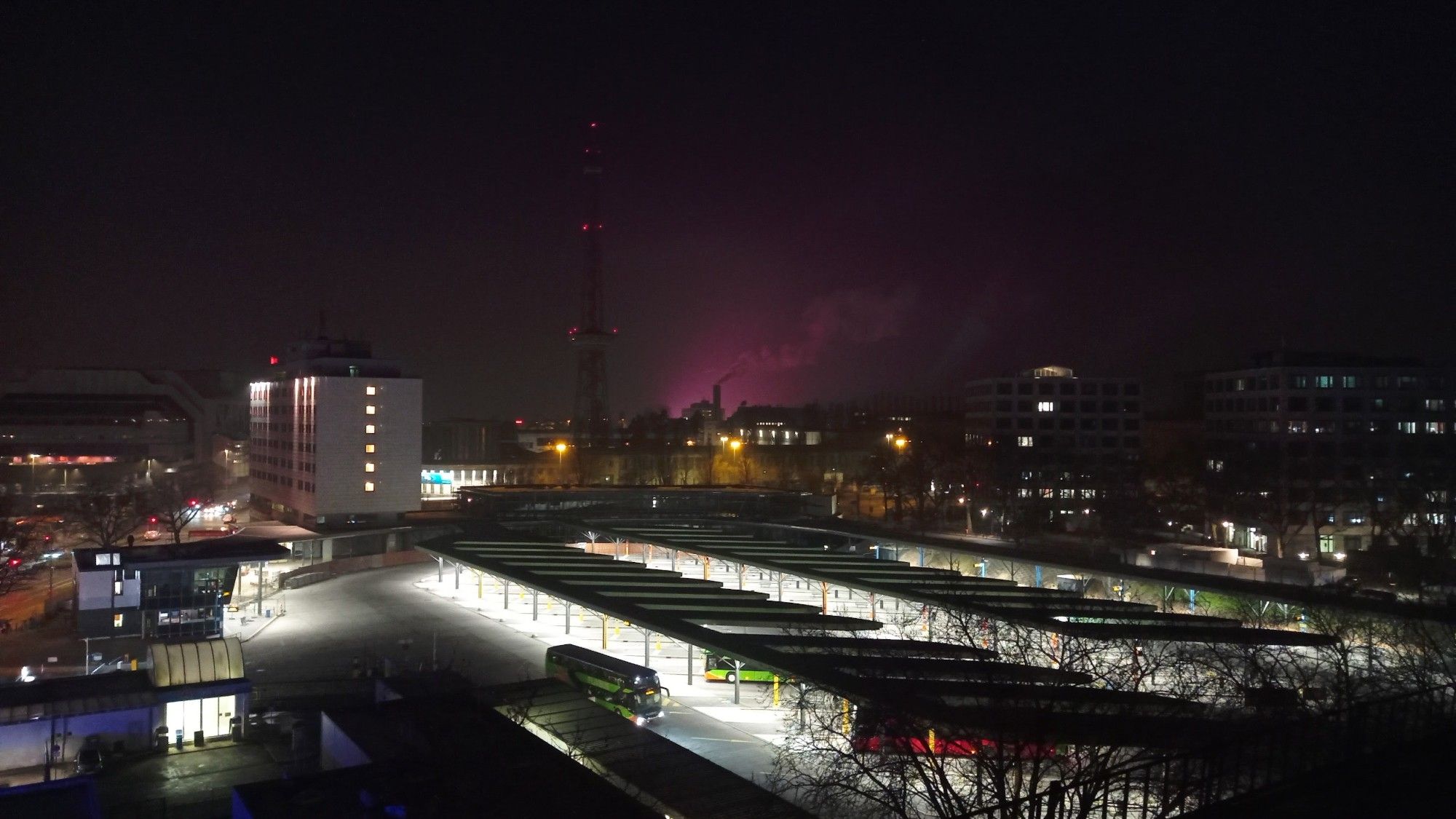 Eine Nachtaufnahme von Charlottenburg in Richtung Zehlendorf. In der Mitte des Bildes ist der Funkturm zu sehen. Rechts vom Funkturm ist lila angestrahlter Rauch oder Dampf zu sehen.