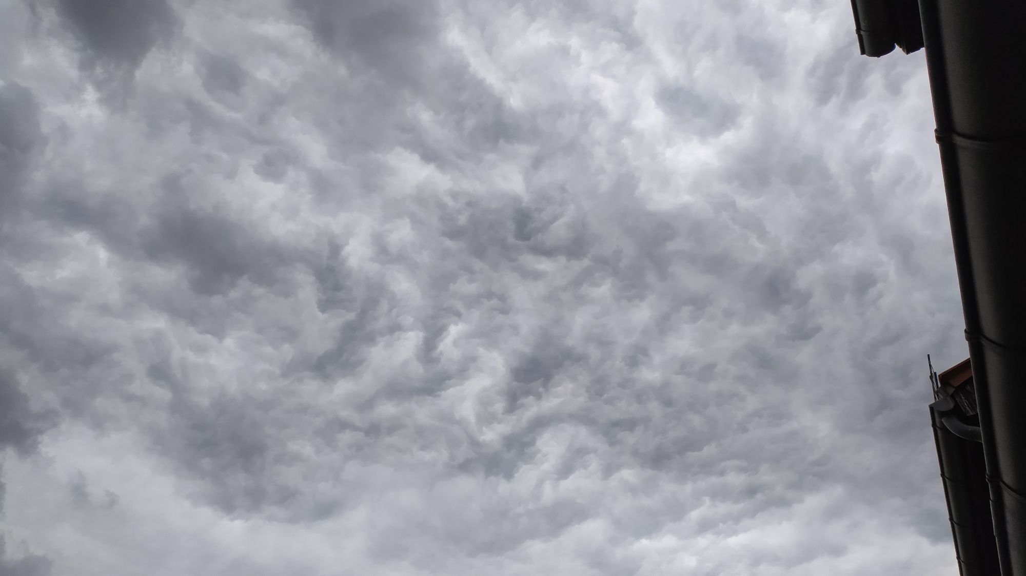 Ein chaotisch anmutendes Muster einer dichten, grauen und dunklen Wolkendecke ist von unten zu sehen. Der rechte Bildrand ist von einer Dachrinne abgeschlossen.