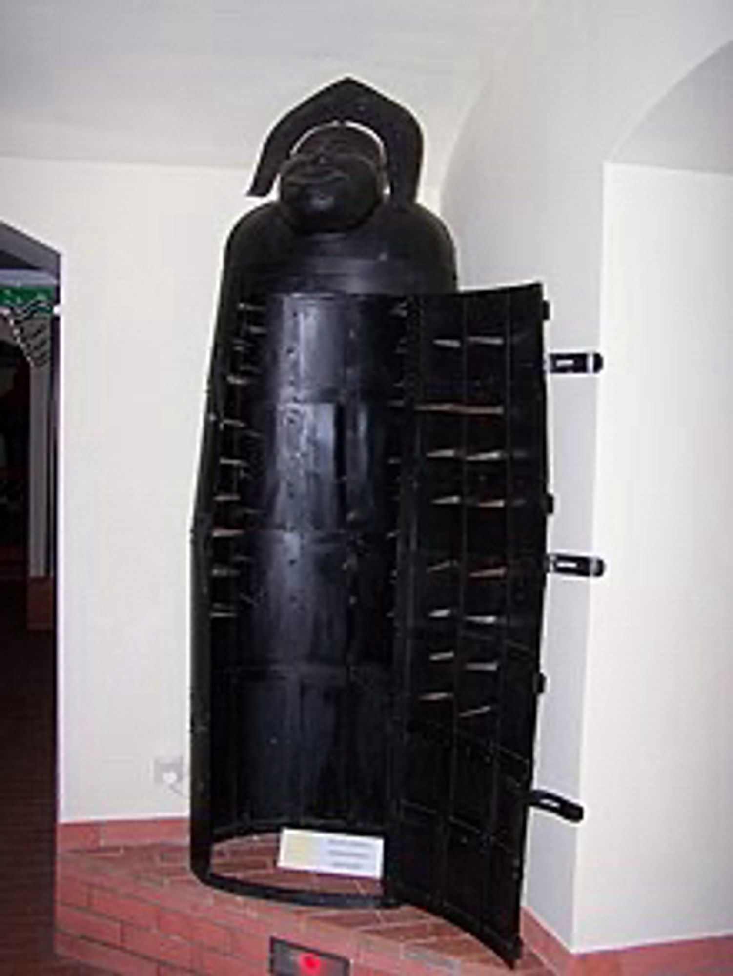 A photo of an iron-maiden-type torture device on display apparently in a museum. It is colored black and covered in interior spikes throughout the torso area.