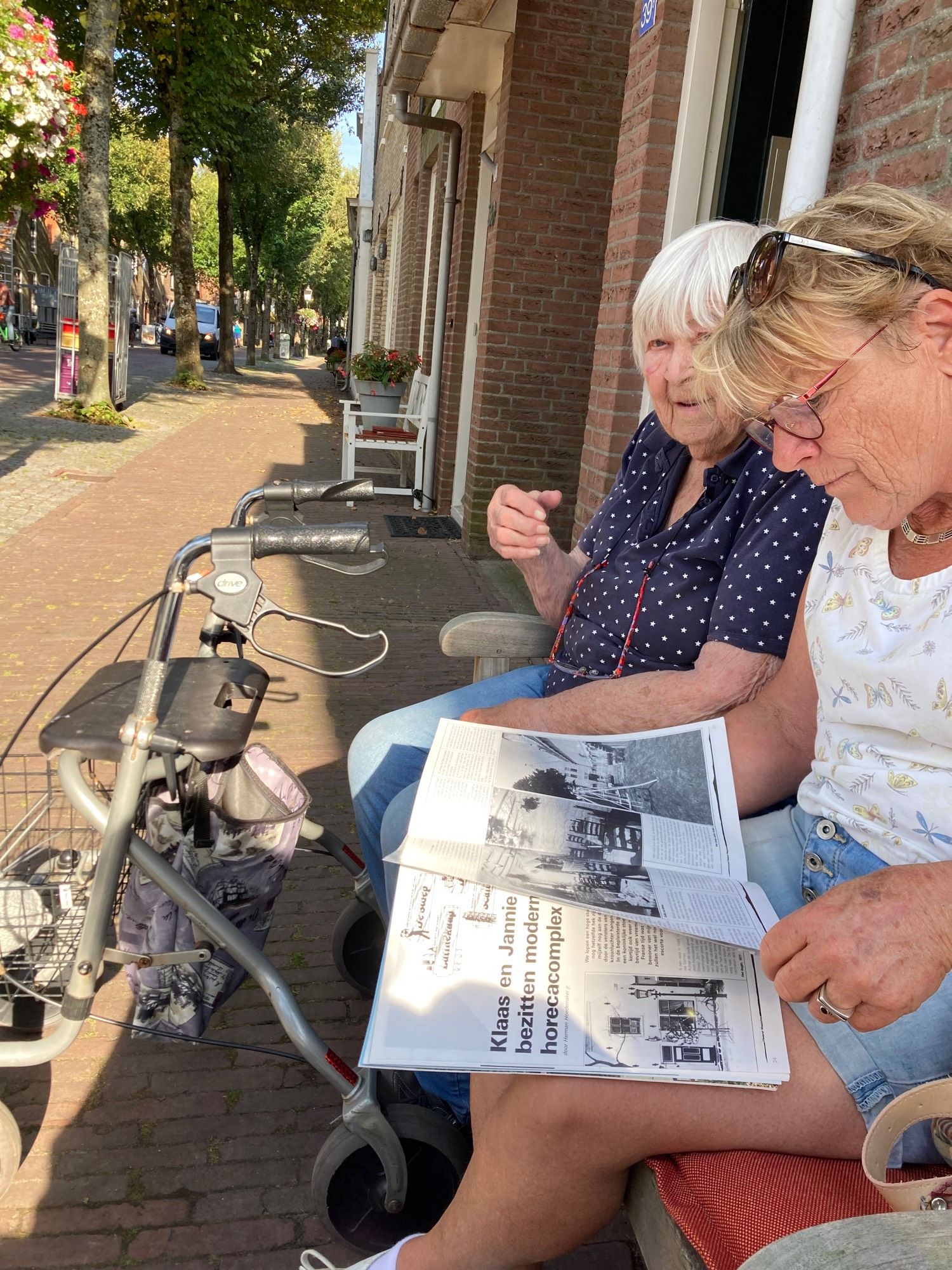 Twee vrouwen op een bankje lezen in een magazine. Eén is in de negentig, de ander rond de zeventig.