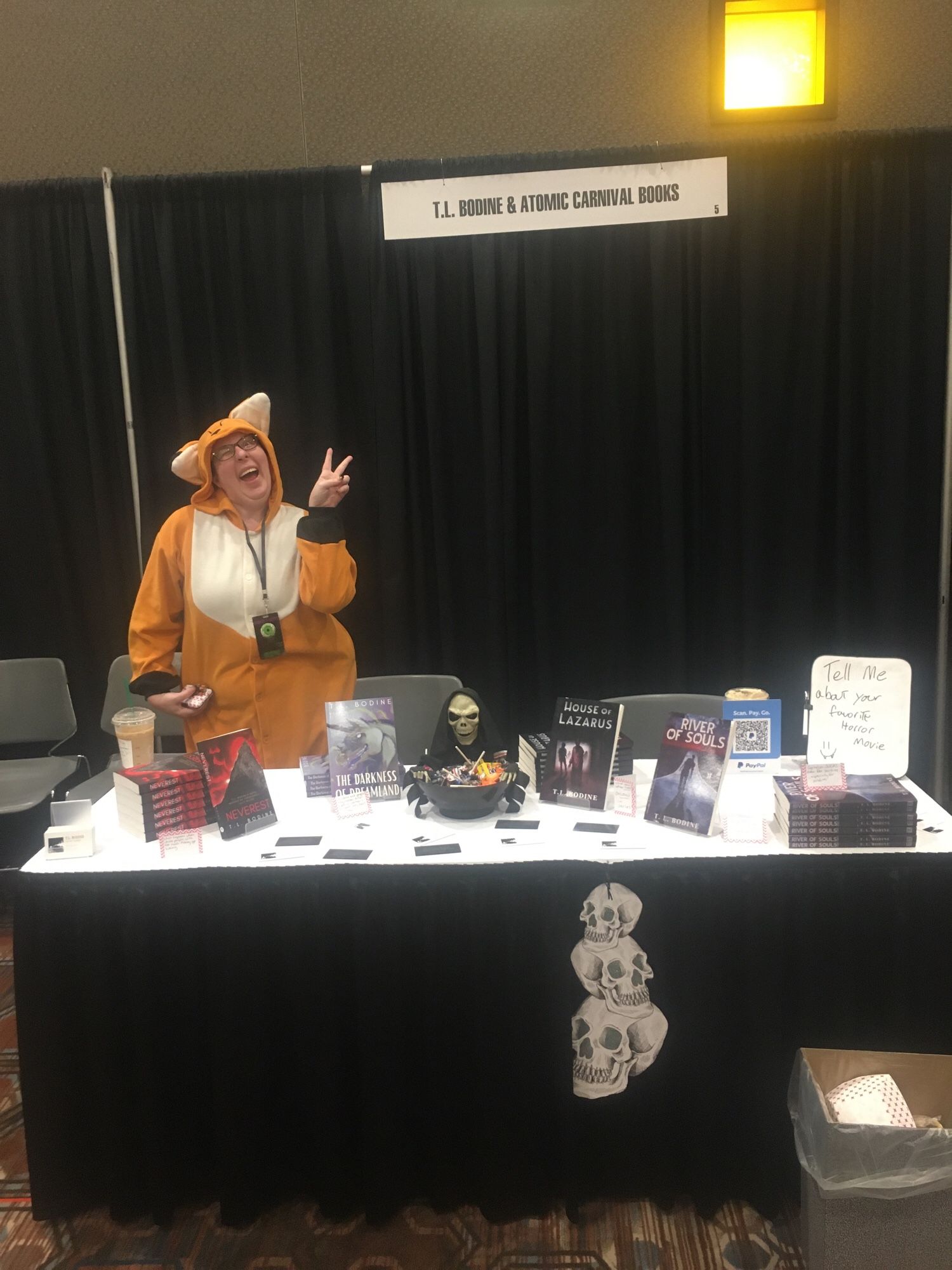 My vendor booth at Morgue & Krypt horror con. There are books and a big bowl of Halloween candy. My booth buddy is wearing conspicuous fox pajamas.