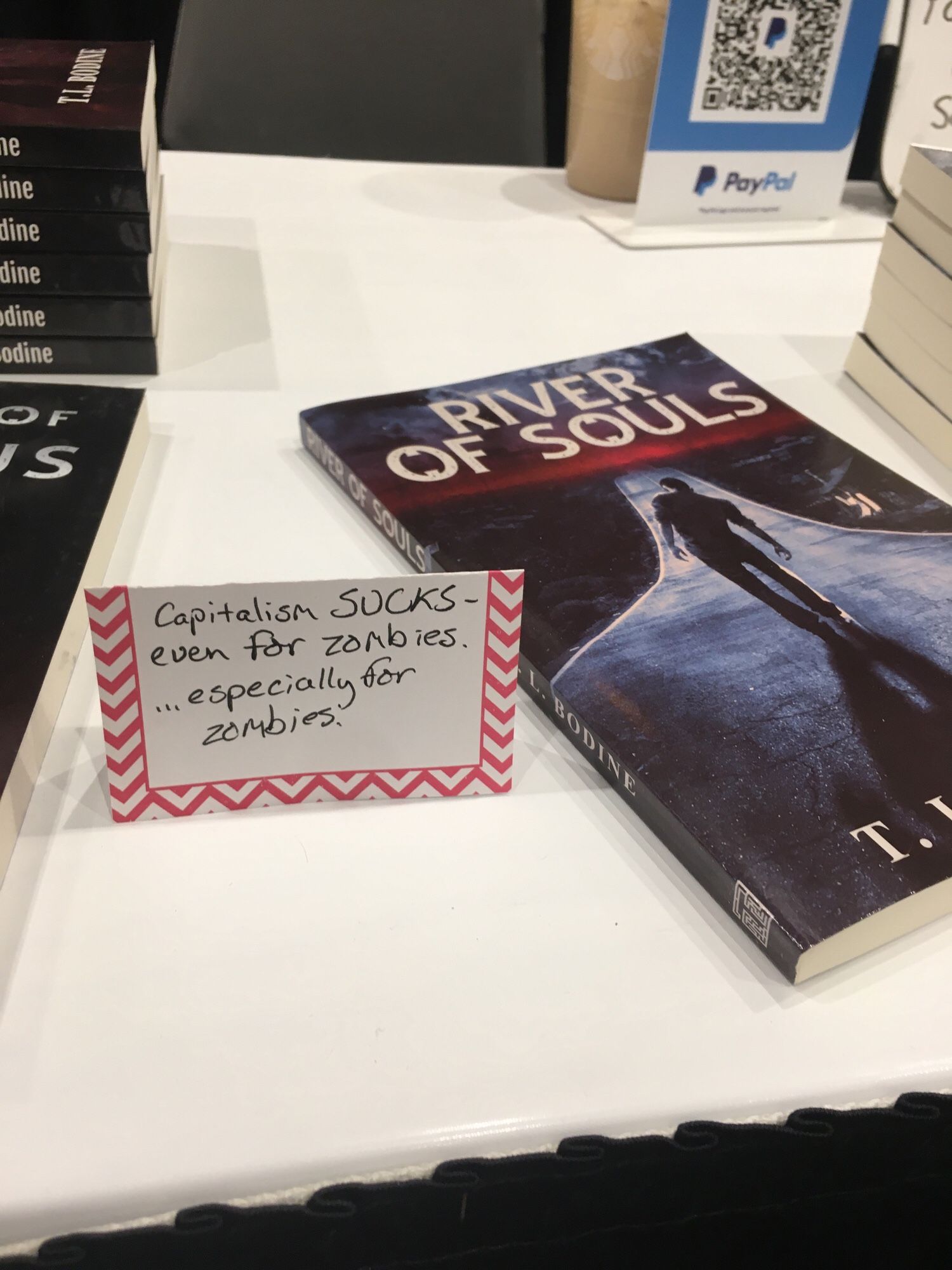 A copy of my novel River of Souls with a sign next to it reading “capitalism sucks even for zombies….especially for zombies”