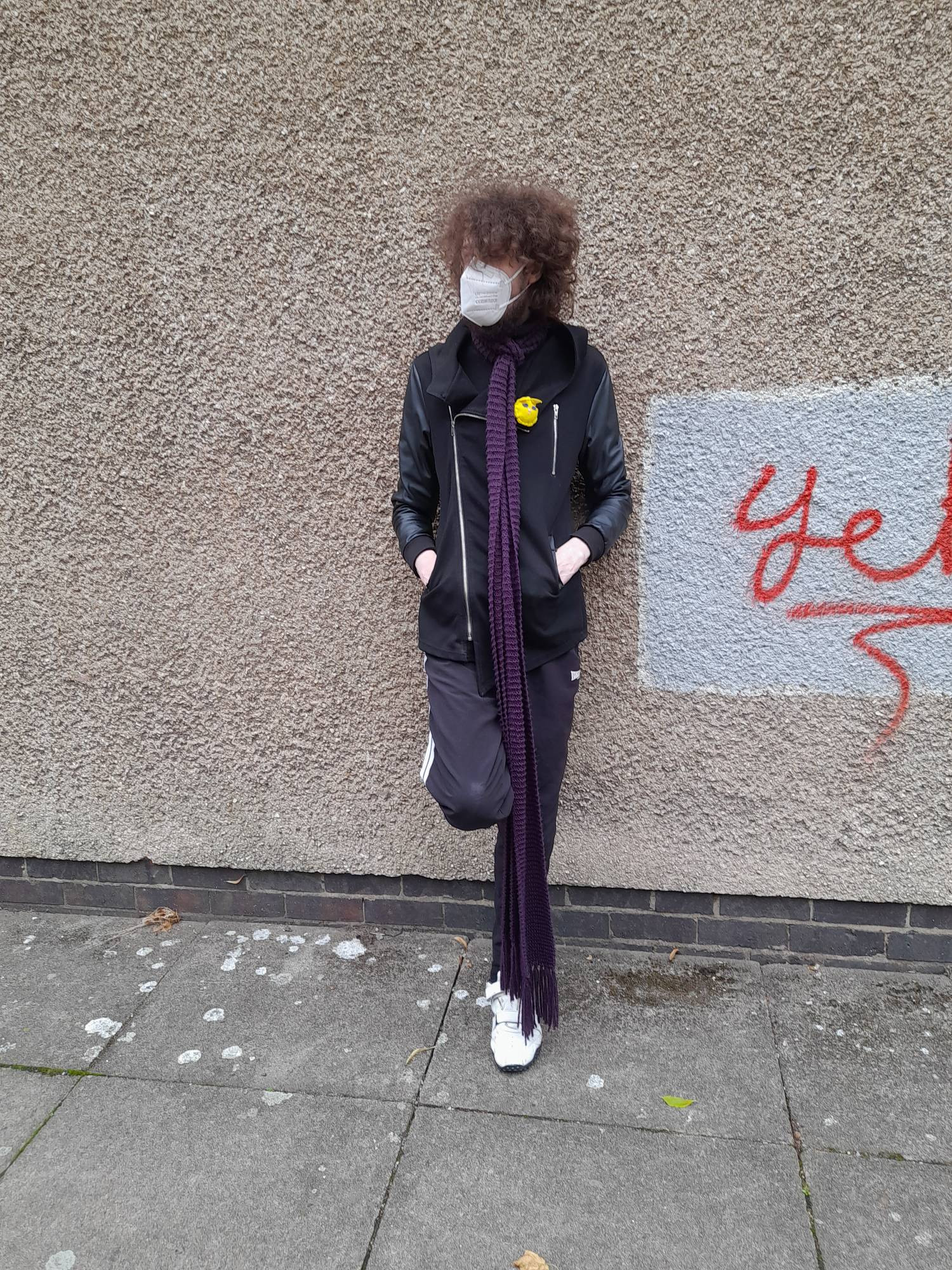 A tall young man with curly hair is standing next to a pebble dashed wall. He is wearing black jogging bottoms with white trainers and a black jacket with an N95 mask and a long burgundy scarf