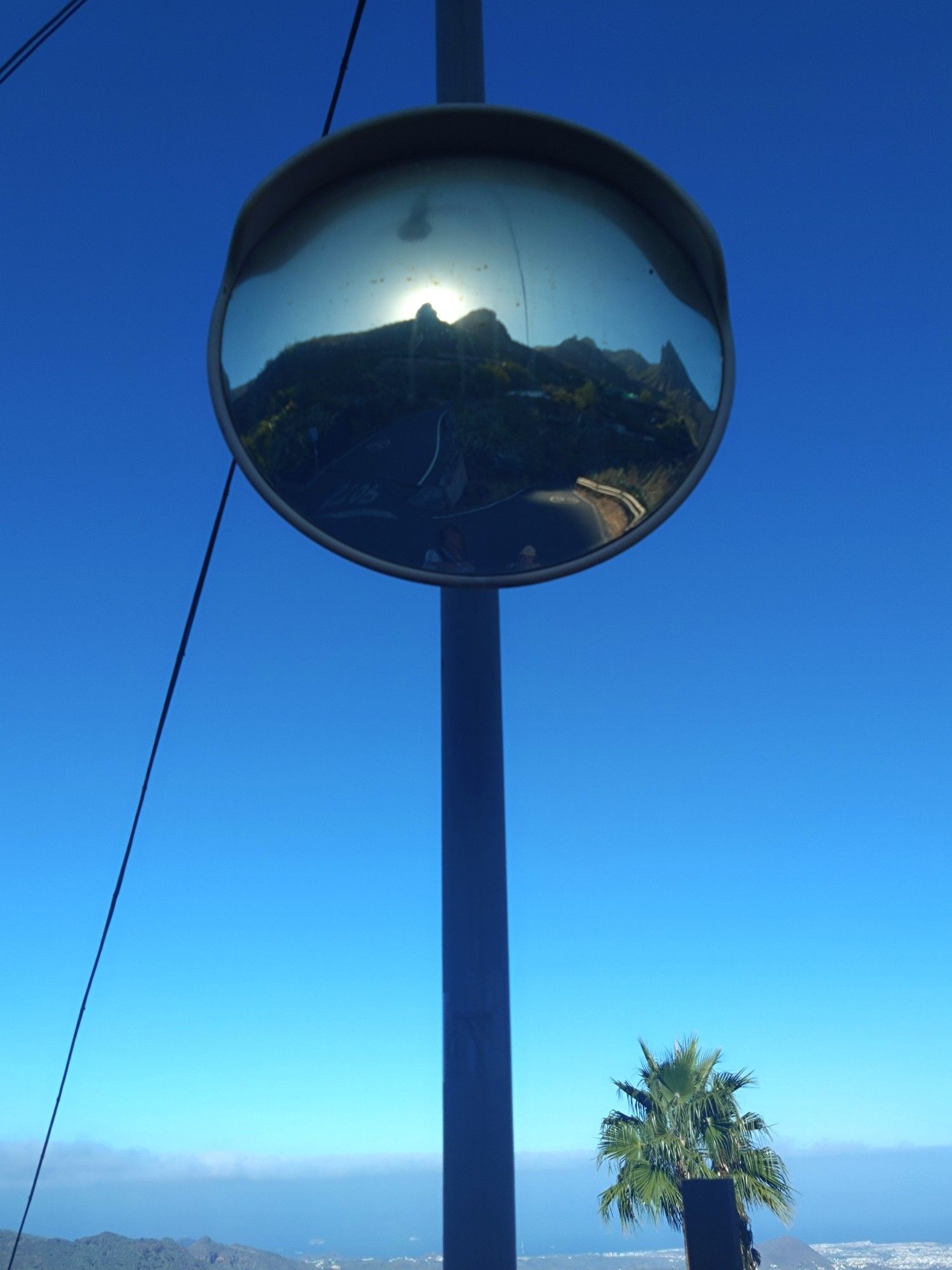 A convex mirror on a post reflects a distorted view of the mountains behind the photographer. The view ahead is of clear blue sky and the top of a palm tree against a backdrop of low cloud and the top of a distant ridge.