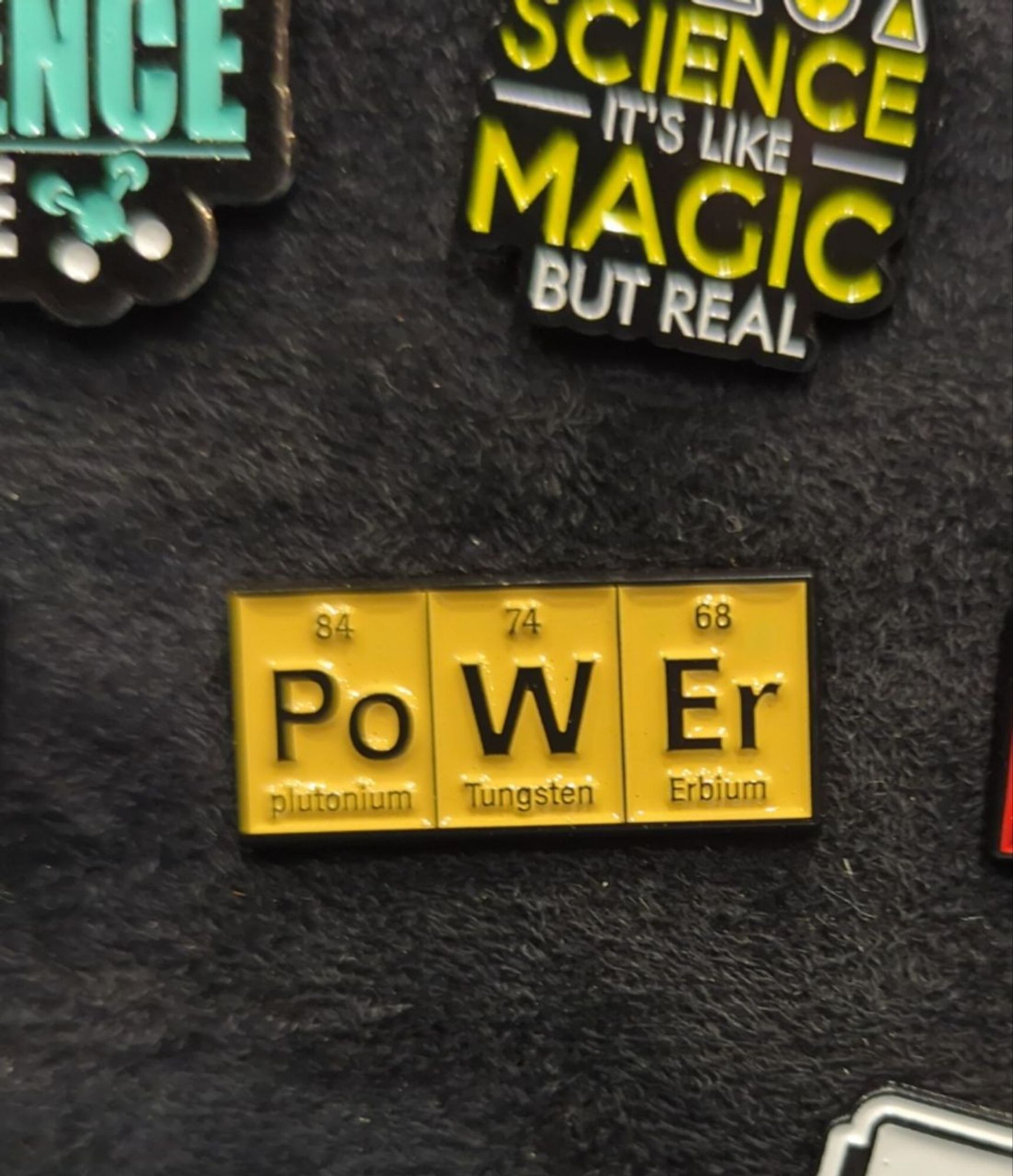 Ein Anstecker mit den Wort »Power« zusammengesetzt aus drei Elementen des Periodensystems. »W« und »Er« sind Wolfram und Erbium, unter »Po« mit Ordnungszahl 84 steht allerdings »Plutonium«.