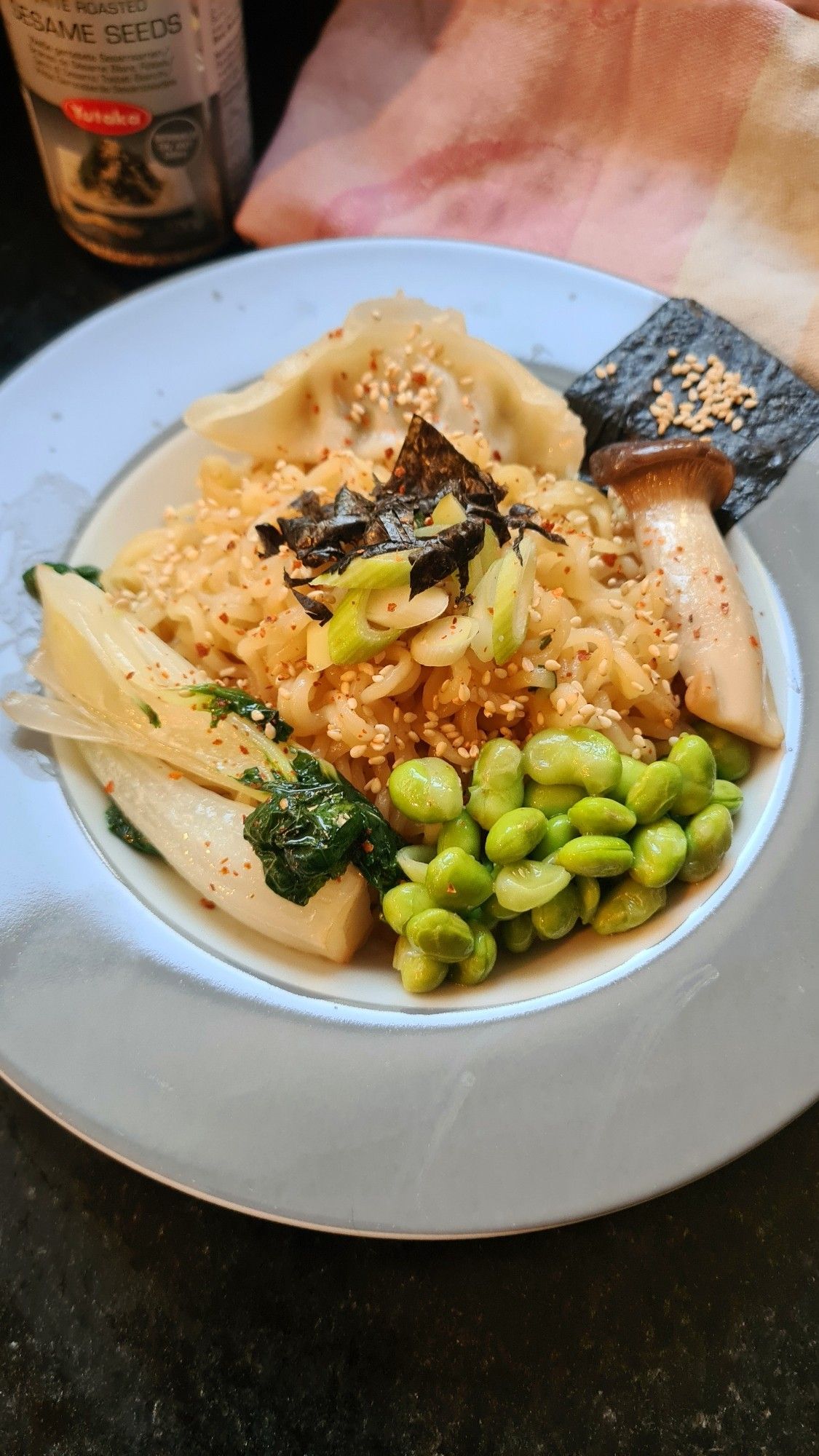 Ramennudeln auf tiefem Teller mit Mini-Pak-Choi, halbem Kräutersaitling, einer Gyoza-Tasche, Edamame, Frühlingszwiebeln, Nori-Blatt-Krümeln und Sesam. Im Hintergrund steht noch der Sedam-Streuer und ein Küchenhandtuch.