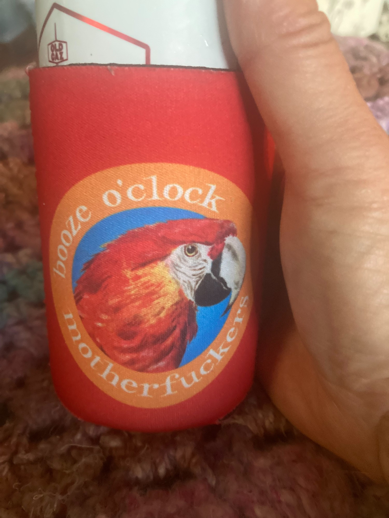 A red can cozy printed with the head of a red and gold macaw. Around that in white letters on an orange circle, the words "booze o'clock motherfuckers."