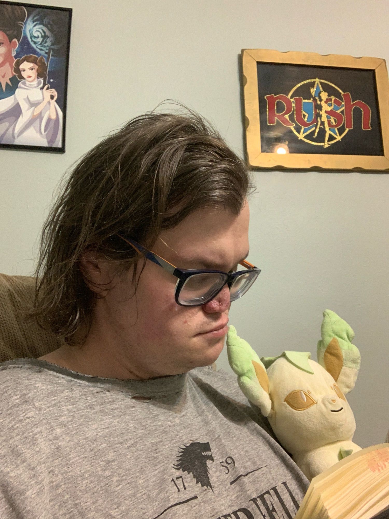 A white person with shortish dark hair and orange and blue glasses sits reading a book. A Leafeon plush is nestled against their left shoulder.